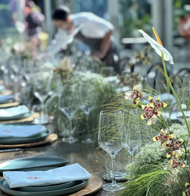 Montaje de mesa elegante con vajilla gris, copas de cristal y centros de mesa con anturios blancos, orquídeas y follaje verde. Al fondo, un camarero ajusta los detalles bajo la luz natural que entra por los ventanales.