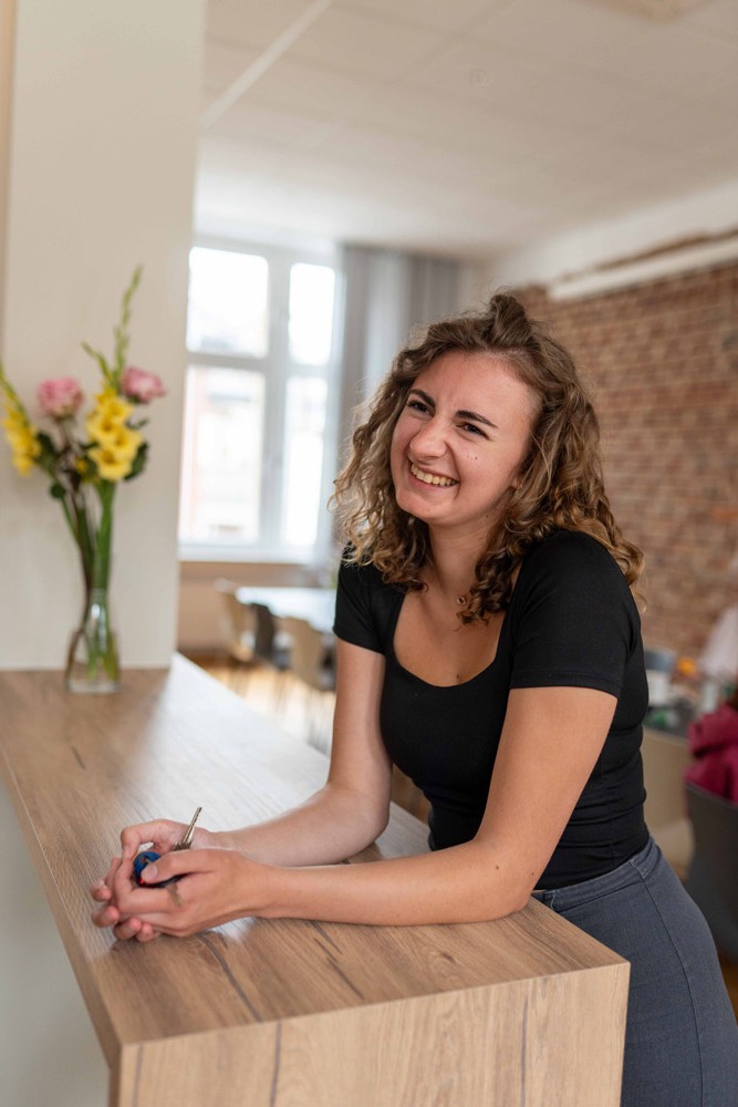 Mitarbeiterin im schwarzen Oberteil sitzt am Schreibtisch, im Vordergrund ein Blumenstrauß, im Hintergrund Backsteinwand.