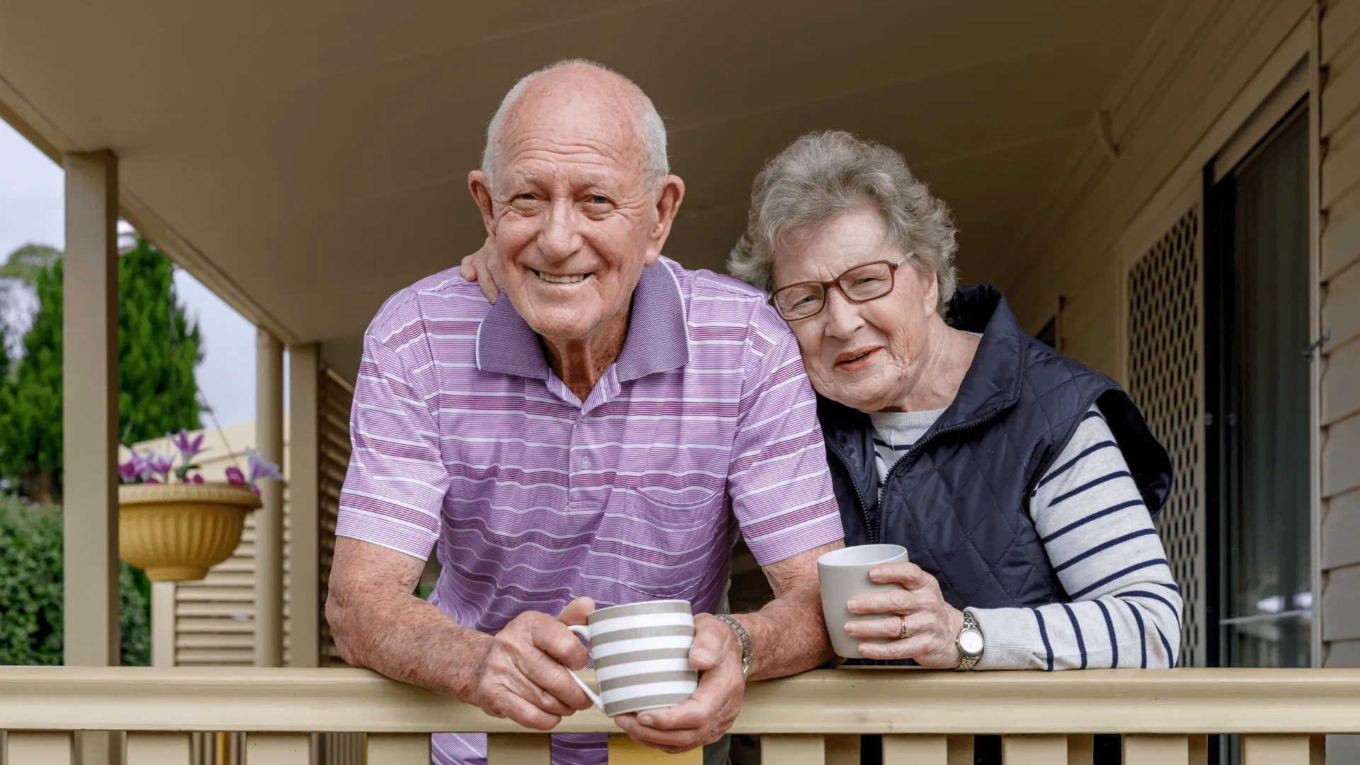 Two elderly people leaning together on a railing, holding mugs