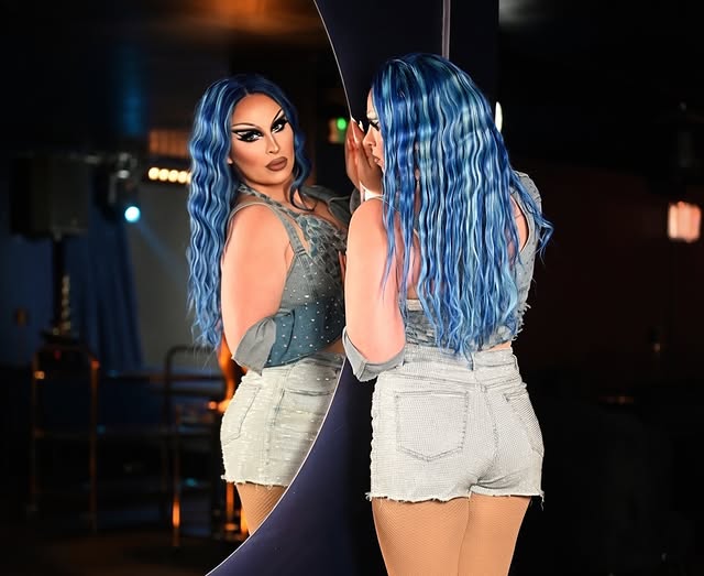 A drag performer with vibrant blue wavy hair and dramatic makeup looking at her reflection in a large mirror, wearing a distressed denim two-piece outfit.