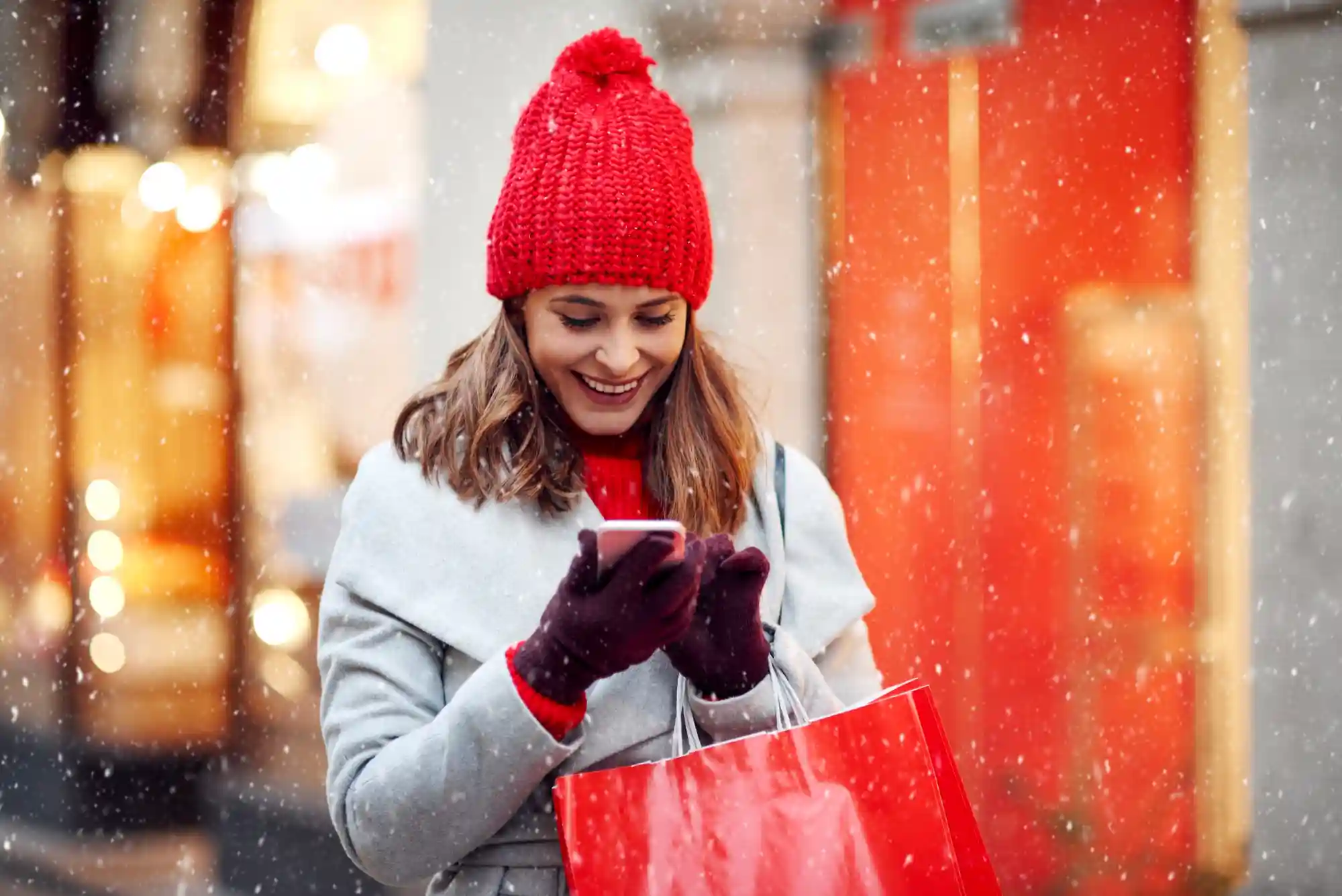 Woman in a red beanie using her phone while holding a shopping bag on a snowy city street.