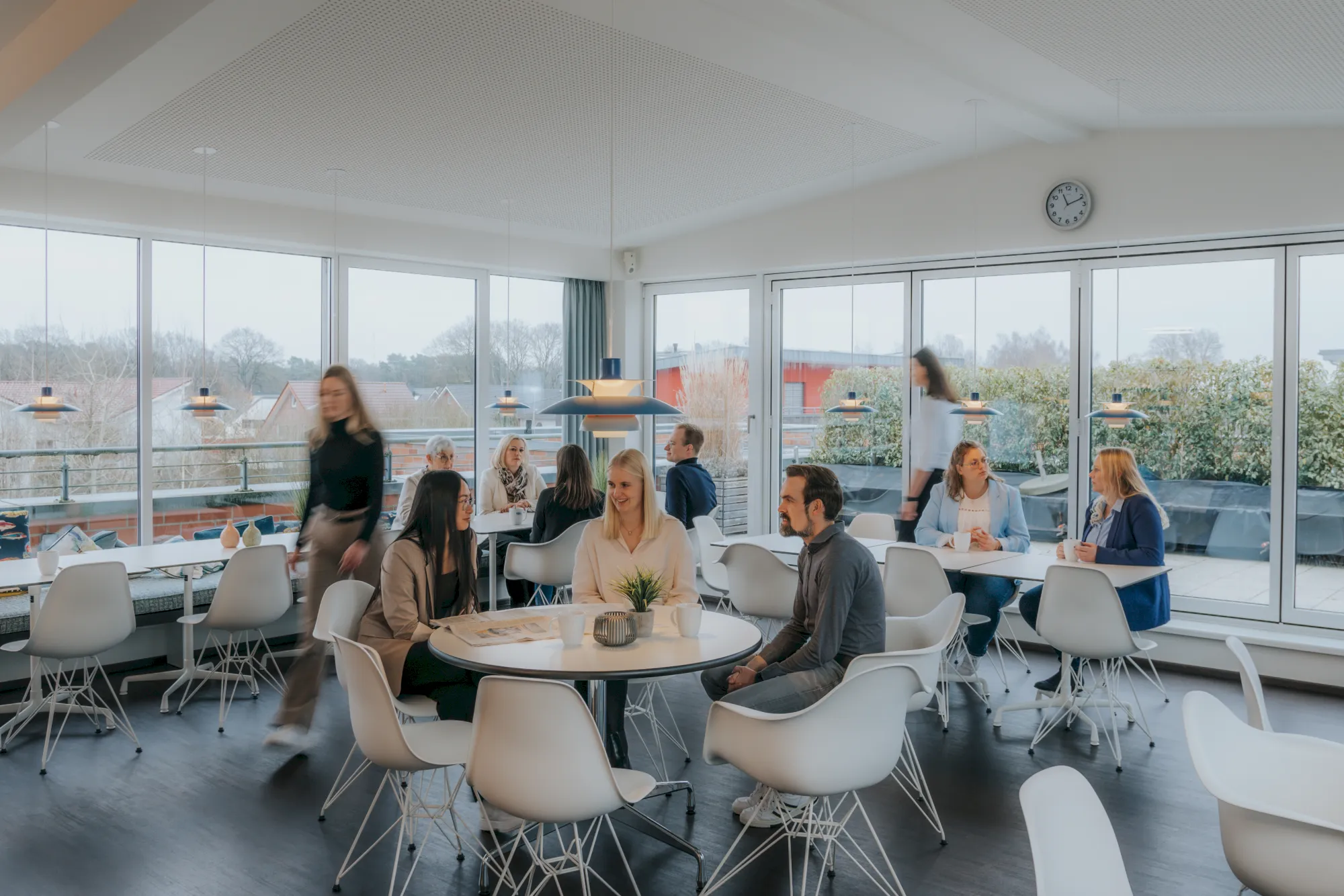 Heller, moderner Pausenraum mit Menschen an Tischen und grossen Fenstern. Entspannung und Kommunikation.