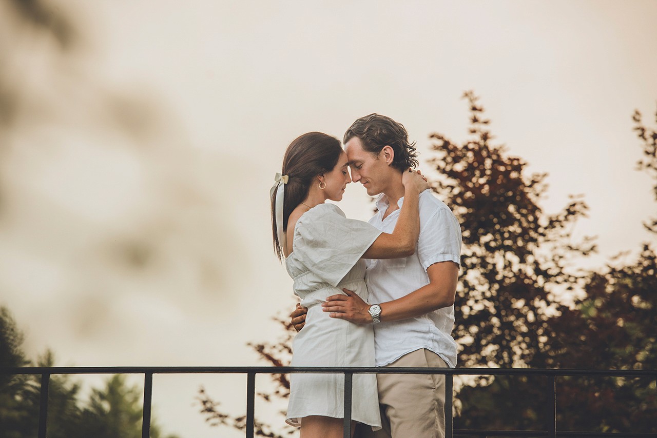 Couple embrace romantically outdoors. Woman in white dress, man in white shirt, standing on balcony with arms around each other.