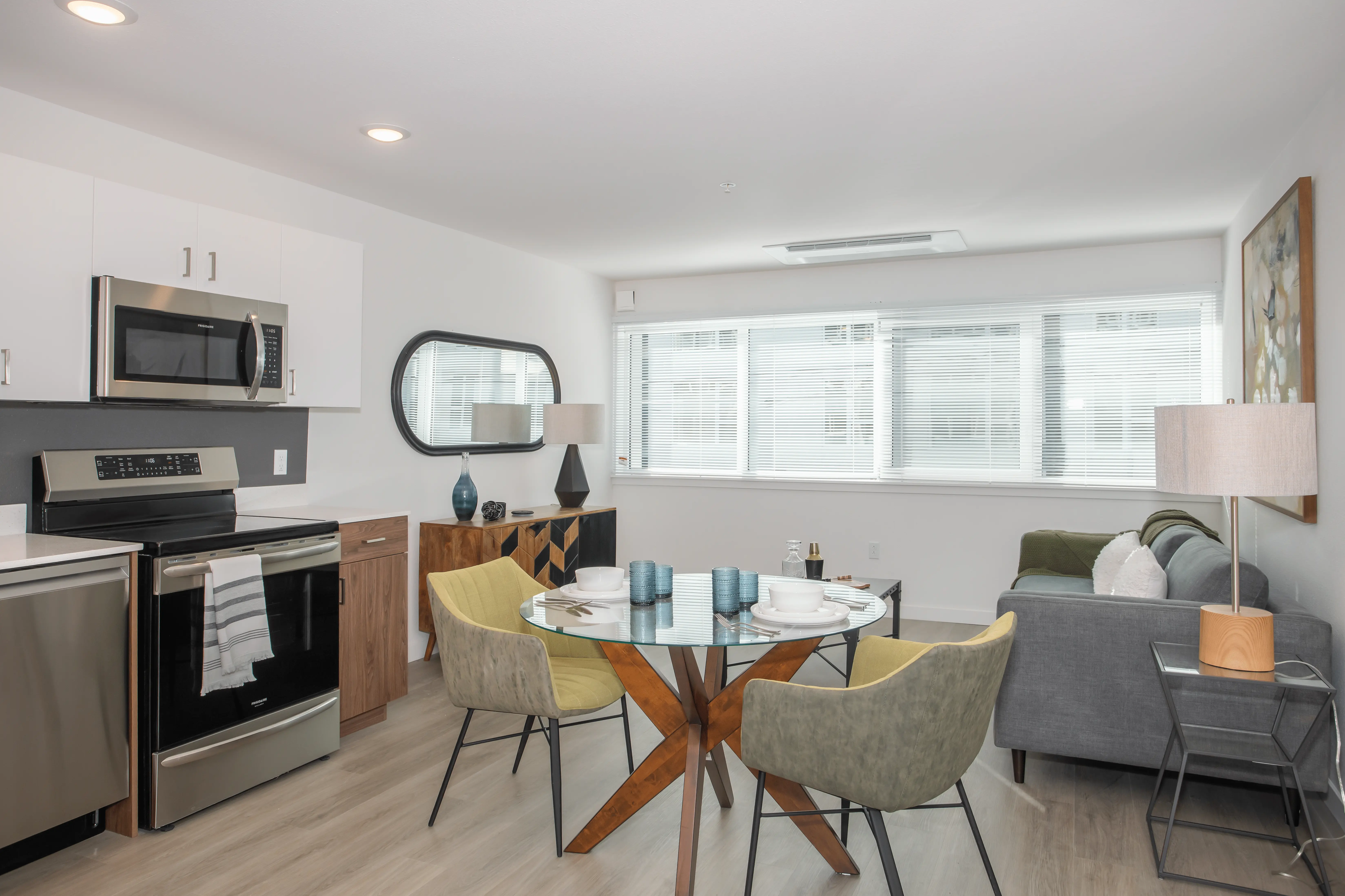 A sun-drenched, modern open-concept living space in Midtown Sacramento featuring a kitchen with stainless steel appliances, white cabinetry, and a black backsplash. Adjacent is a dining area with a round glass table and four chairs. The living area has a gray sofa, a lamp, and a large window.