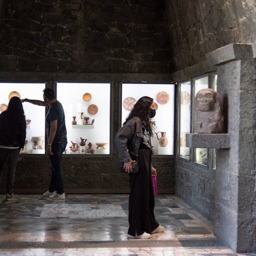 Mga tao na nagmamasid sa sinaunang palayok at mga artifact na naka-display sa isang medyo madilim na museo na may pader na bato.