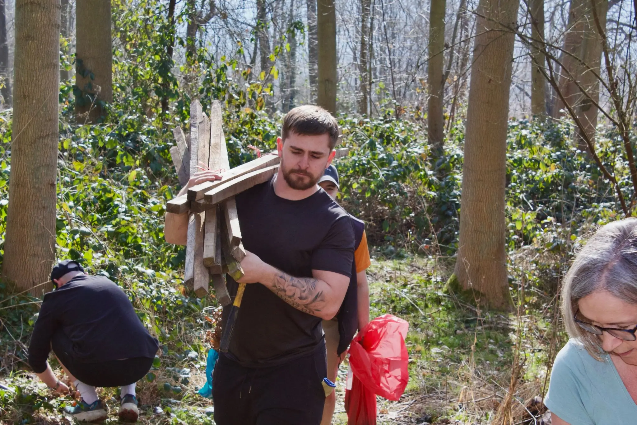 A person stands in a forest, holding an object and smiling, with others working in the background.