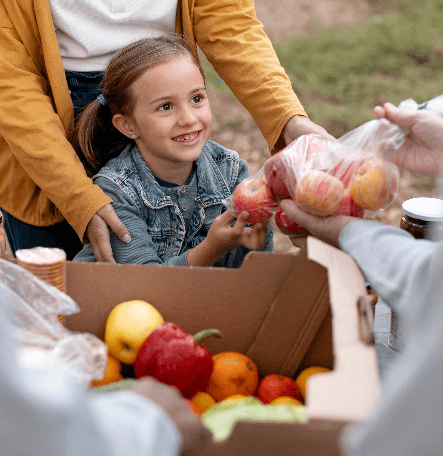 A child smiling while receiving fruits from adults, with a box of assorted fruits in the foreground.