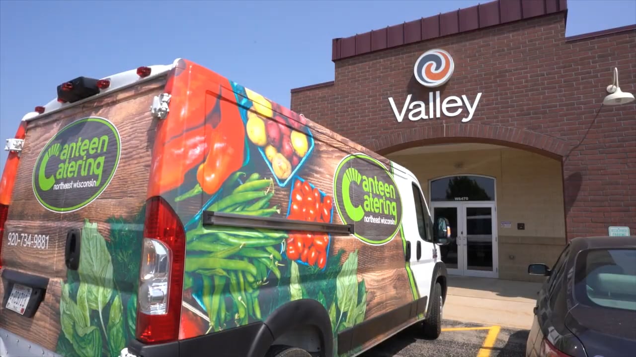 A branded delivery van featuring vibrant images of fresh vegetables is parked outside a brick building with a sign reading "Valley" on a clear day.