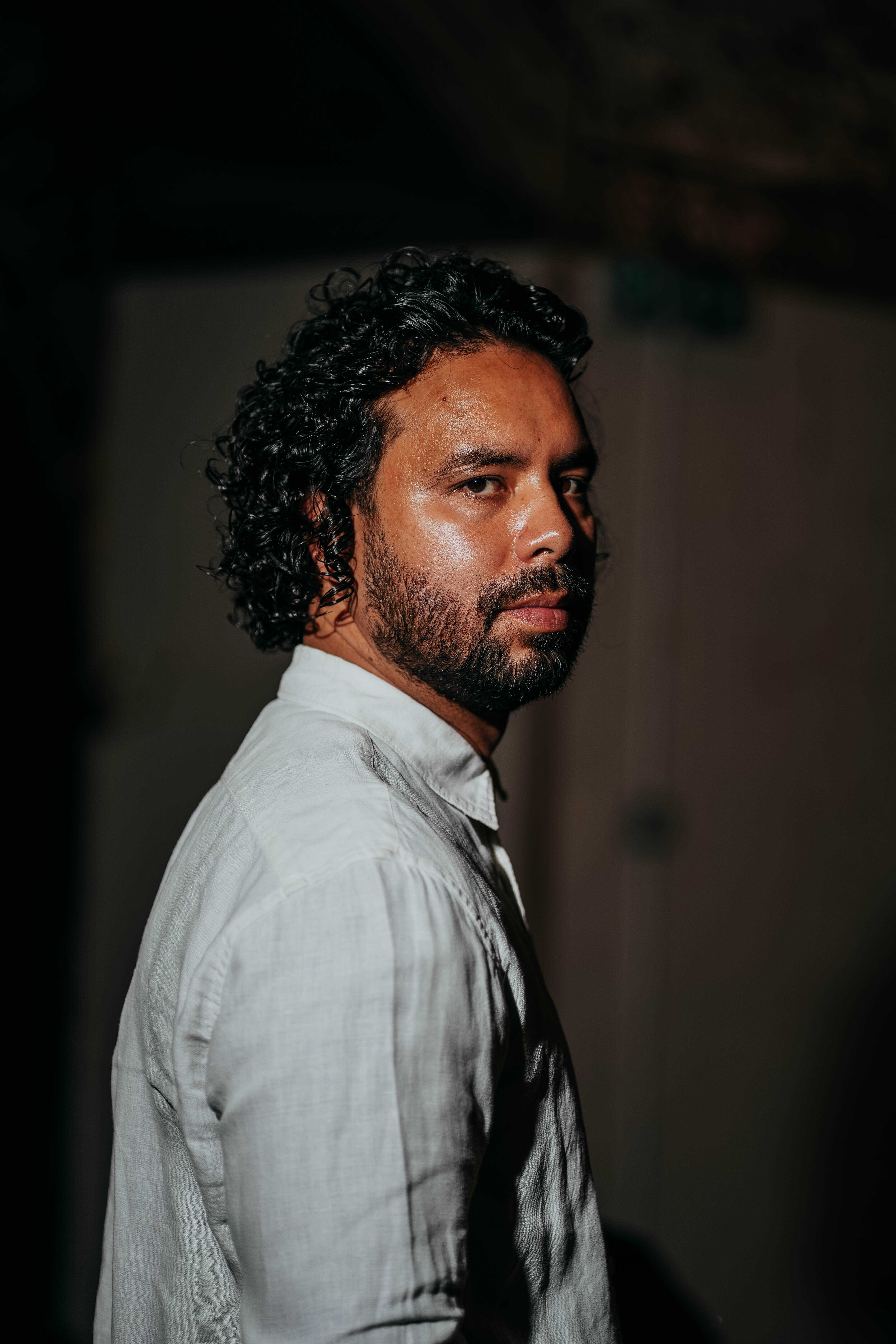 A man with dark curly hair and a beard, wearing a white button-down shirt, stands looking directly at the camera in a dimly lit room with a large, out-of-focus stained-glass window behind him.