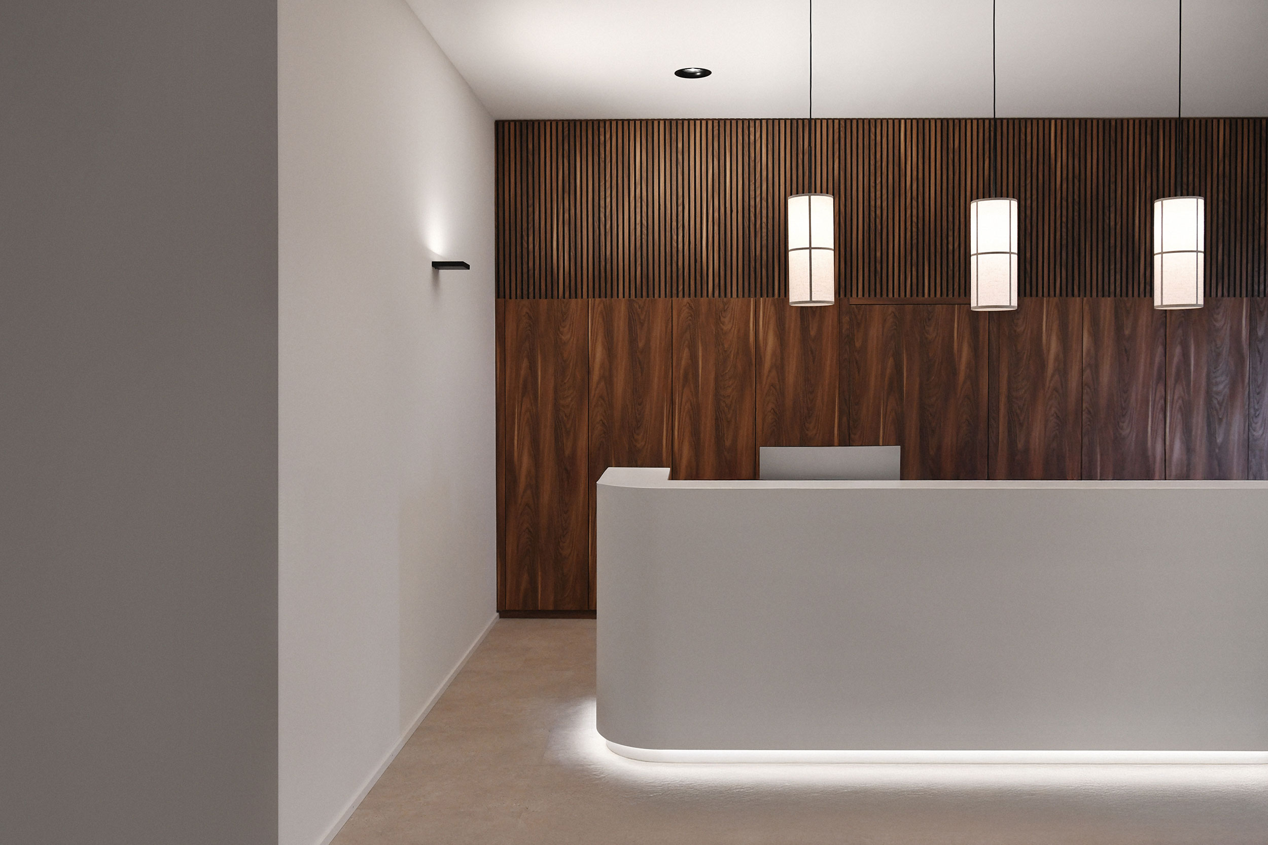 Empfangsbereich der Praxis Spinamed in Muri mit weißer Rezeptionstheke, hölzerner Lamellenwand und drei Pendelleuchten.