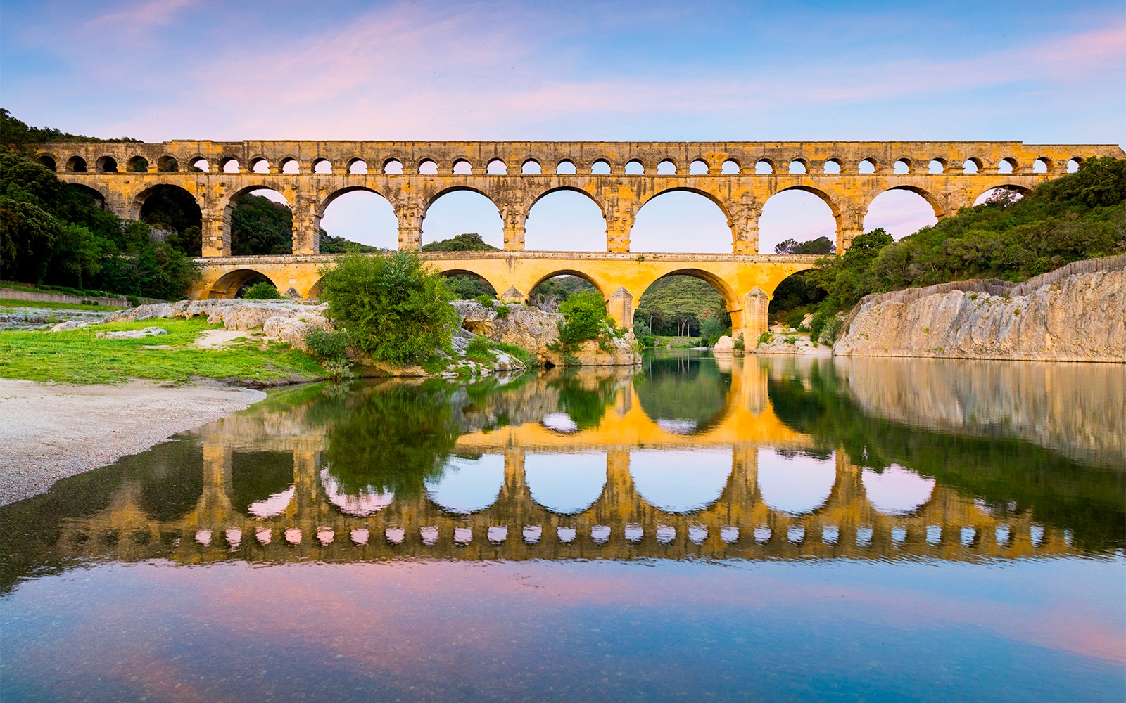 Pont du Gard aqueduct reflecting in the Gardon River, France, with surrounding greenery.