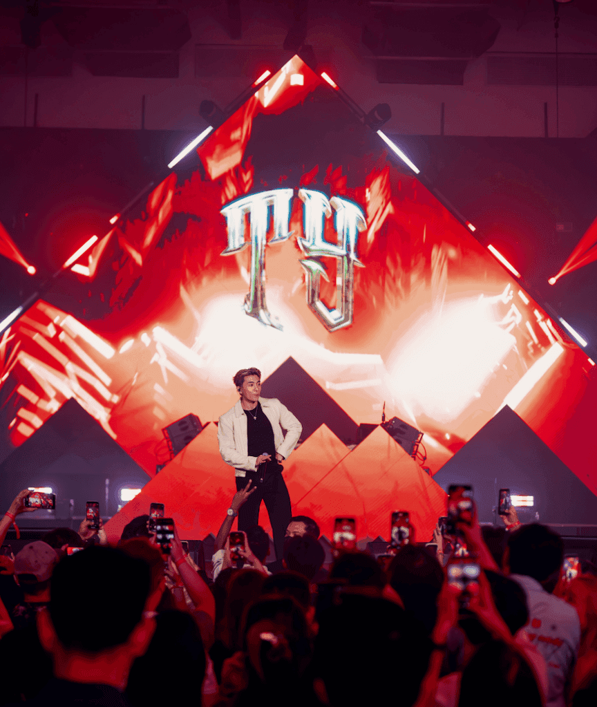 A male performer on stage with a dynamic backdrop at the Hennessy Artistry event, surrounded by an engaged audience holding smartphones.