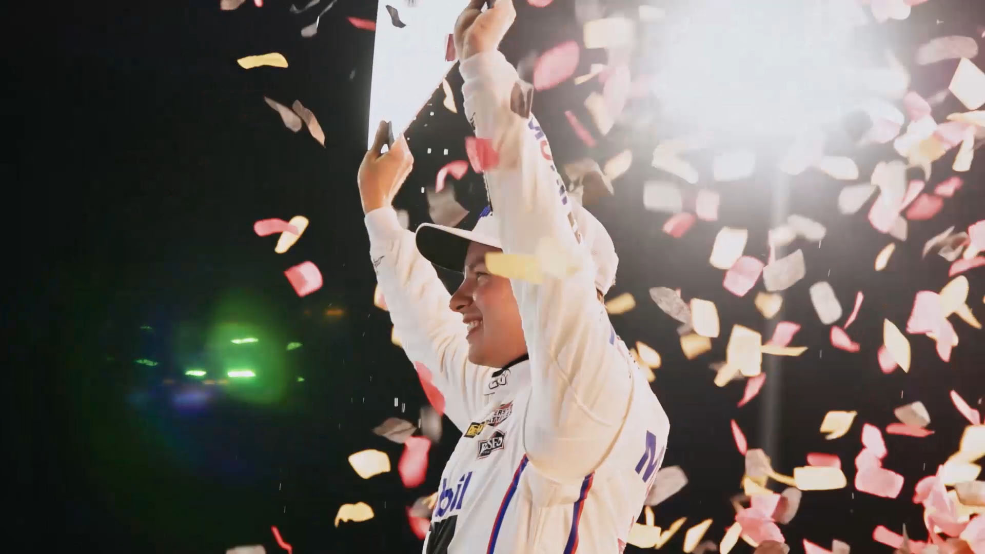 Mobil 1–branded NASCAR driver celebrates on stage, raising a trophy overhead as red and white confetti falls under bright lights.