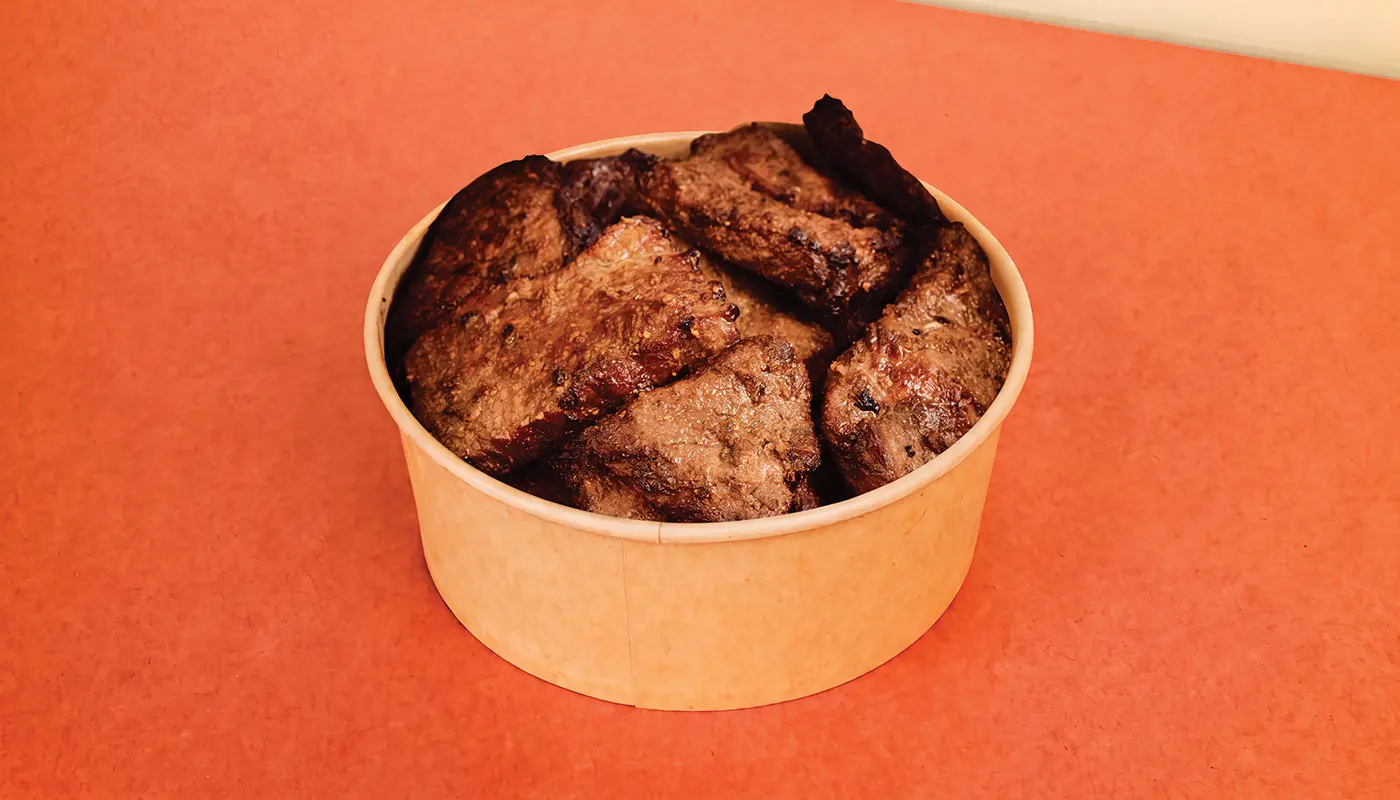 A kraft-paper bowl filled with browned grilled steak pieces with dark charred edges.
