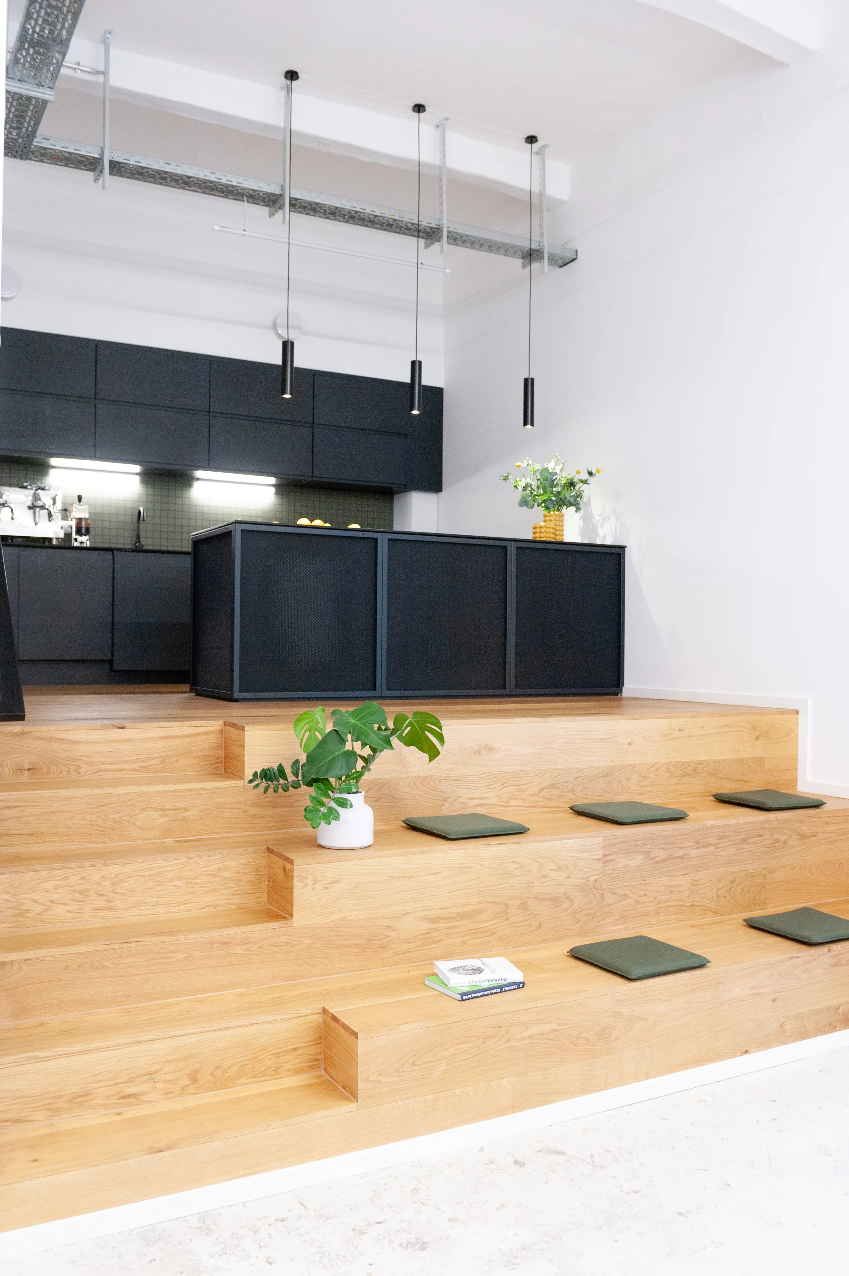 Oak seating tribune with olive-green cushions and potted plants at LA RED, leading up to a matte black kitchen with island counter and slim black pendant lights.