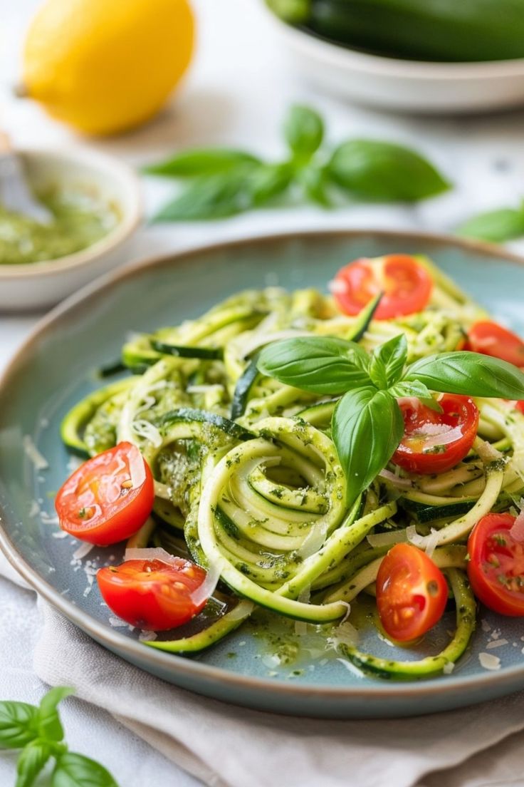 A plate of zucchini noodles tossed in pesto, topped with fresh cherry tomatoes and basil leaves, served on a rustic ceramic plate with lemon and pesto in the background