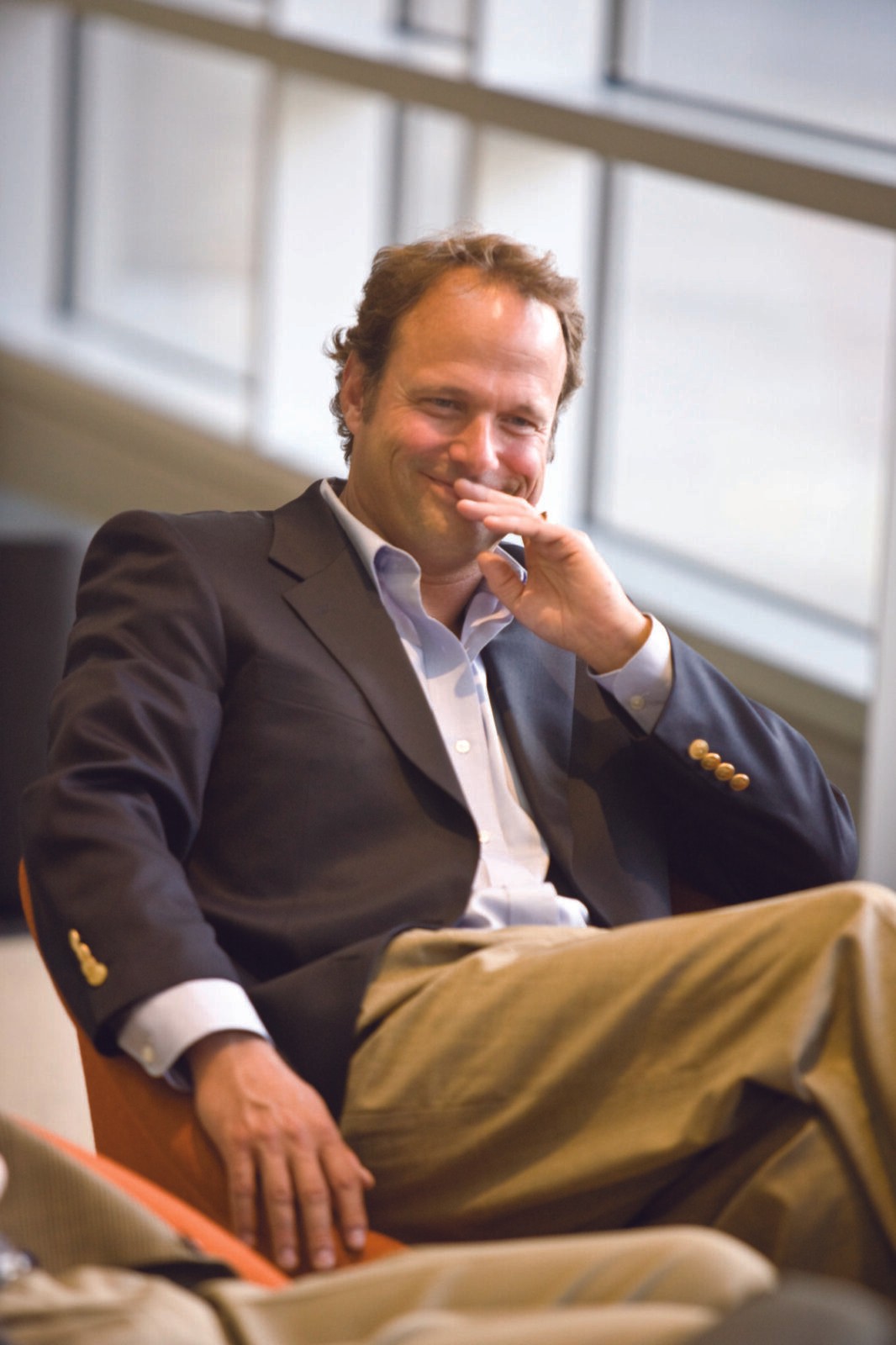 A man in a blazer sits comfortably in a modern office setting, smiling and resting his chin on his hand.
