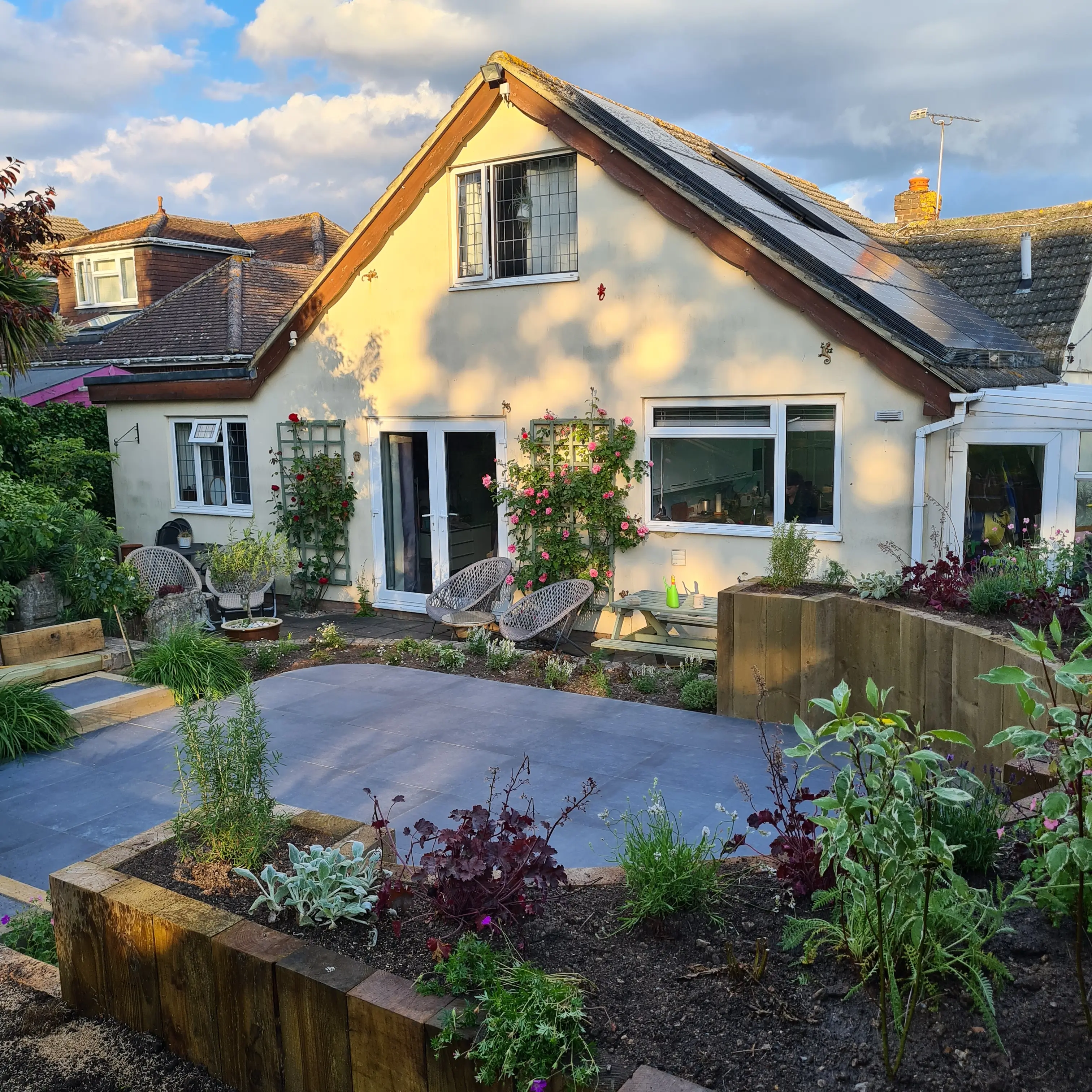 A cozy house surrounded by a vibrant garden, featuring pathways and various plants under a partly cloudy sky.