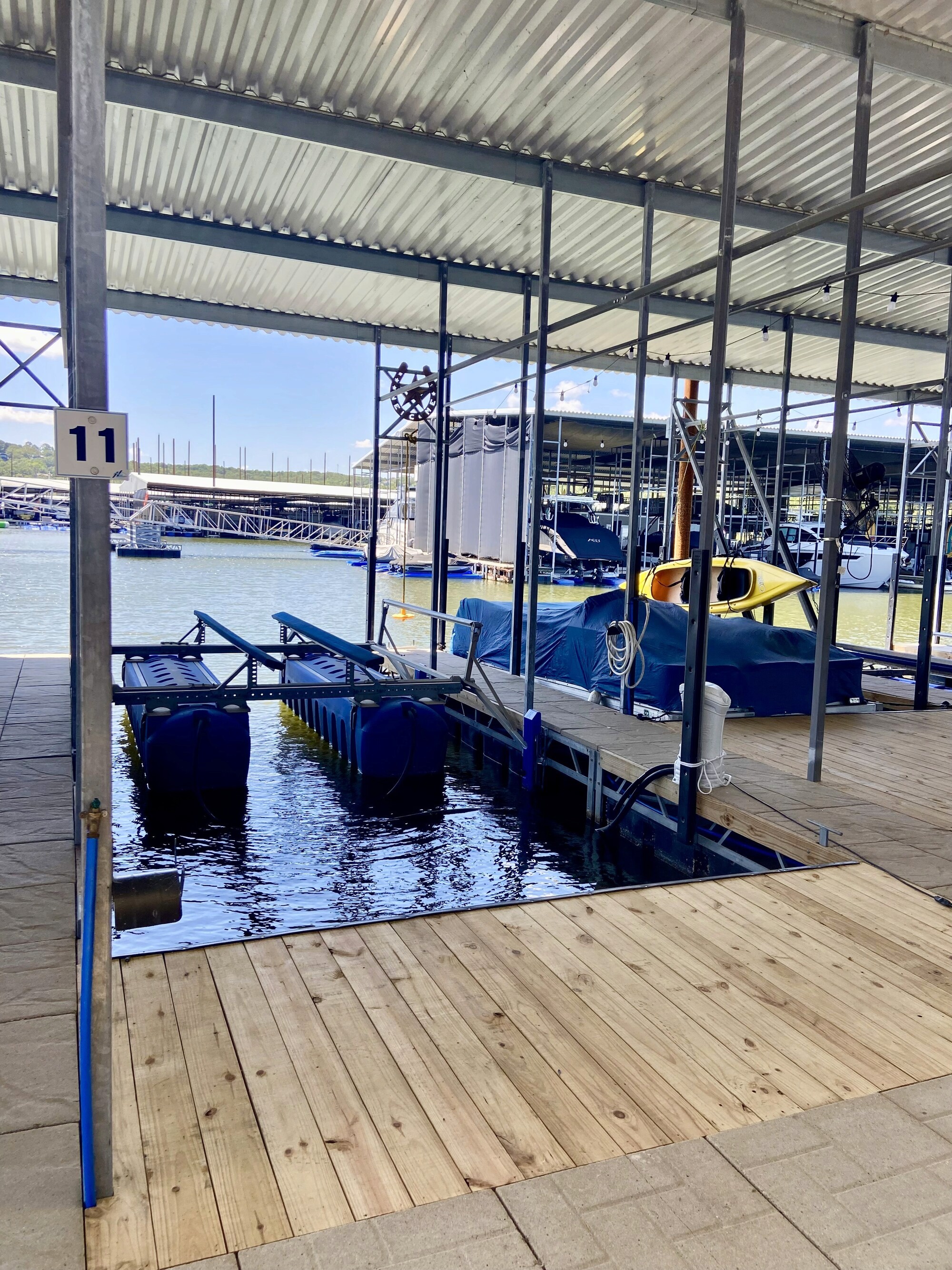 A covered boat slip on a sunny day features a dock with blue barriers and a wooden walkway, overlooking a calm marina with multiple boats and a distant view of a hilly landscape.