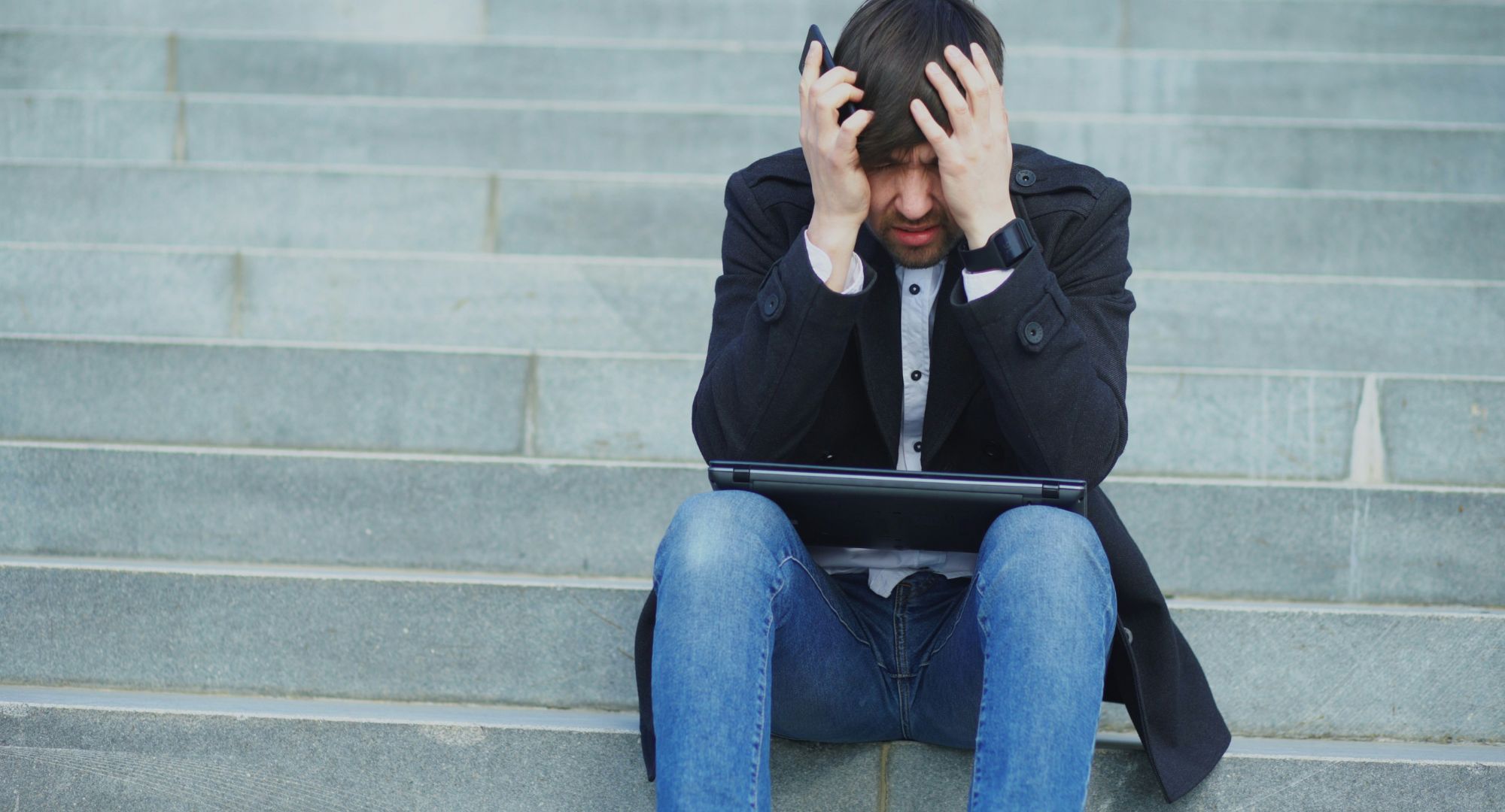 homem sentado em degraus com notebook no colo e as mãos na cabeça frustado, enquanto segura o celular