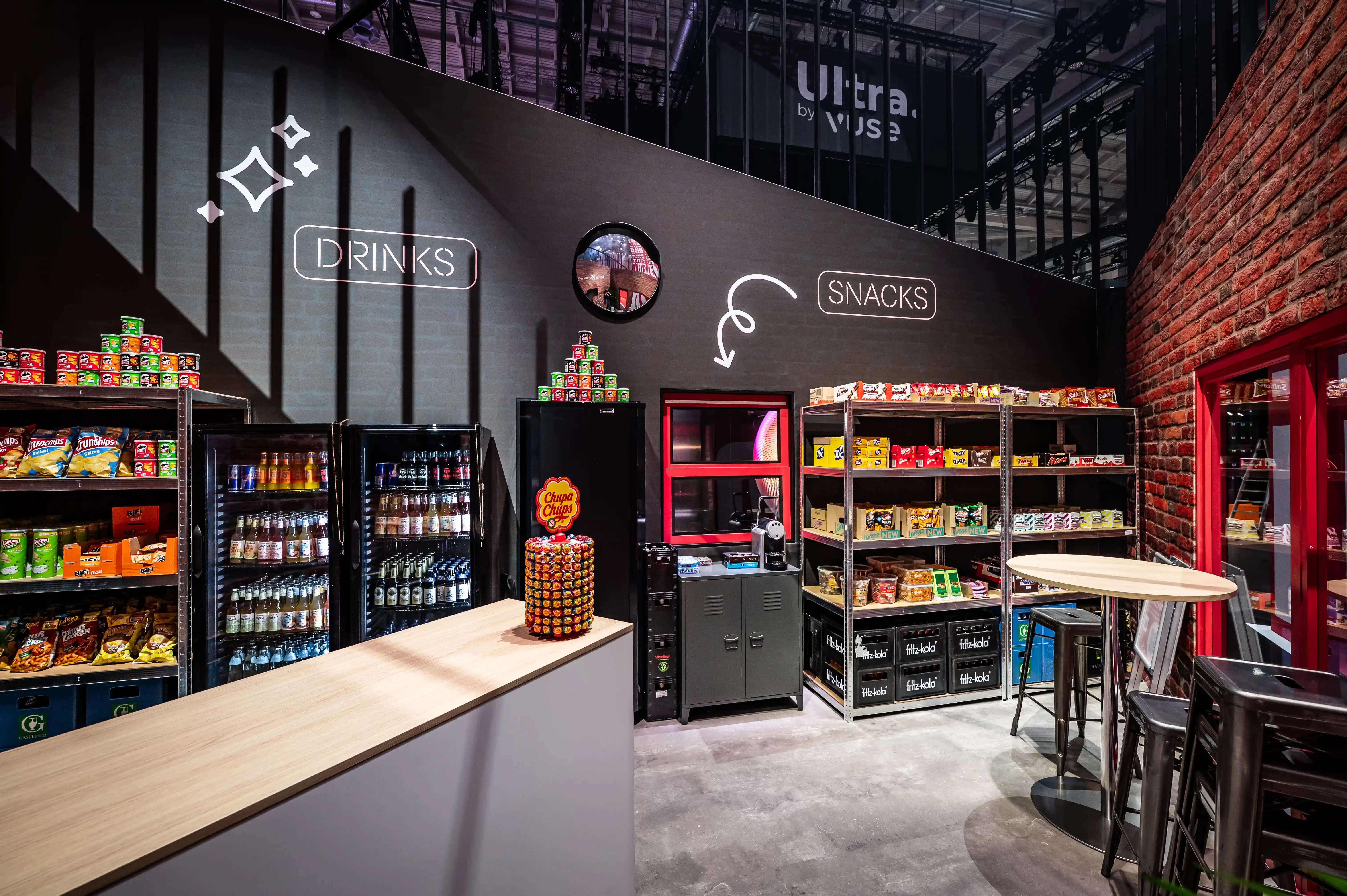 Späti-style kiosk inside the booth with snack and drink shelves, fridges and neon signs on dark walls.
