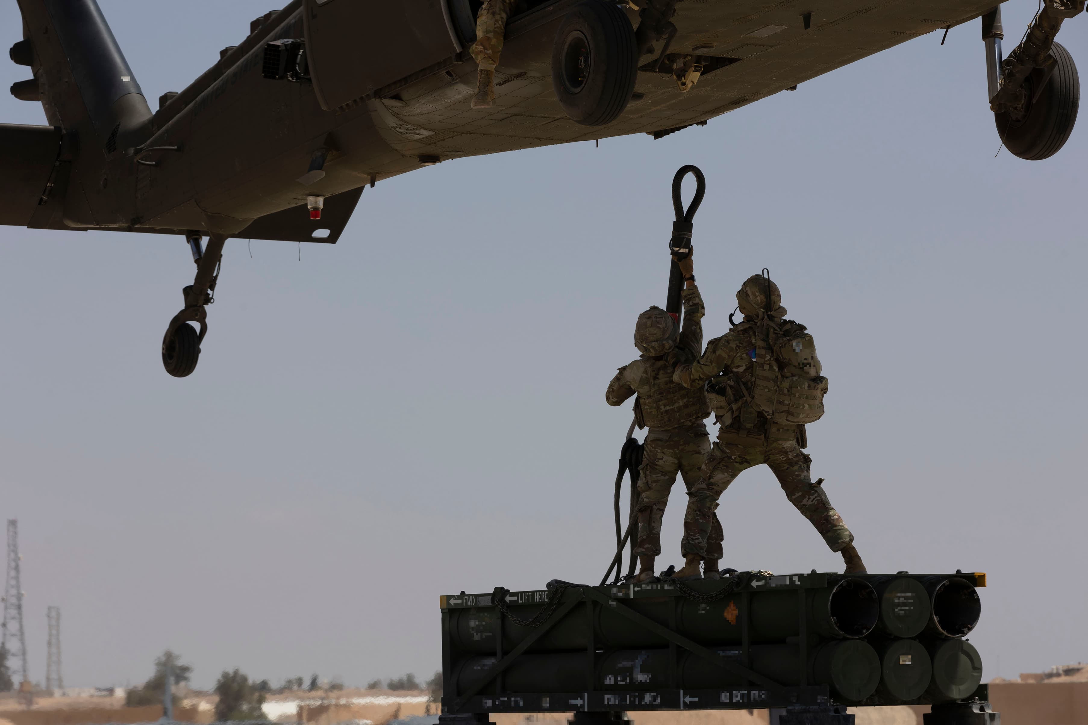 Soldiers in tactical gear securing a sling load to a hovering military helicopter during a training exercise.