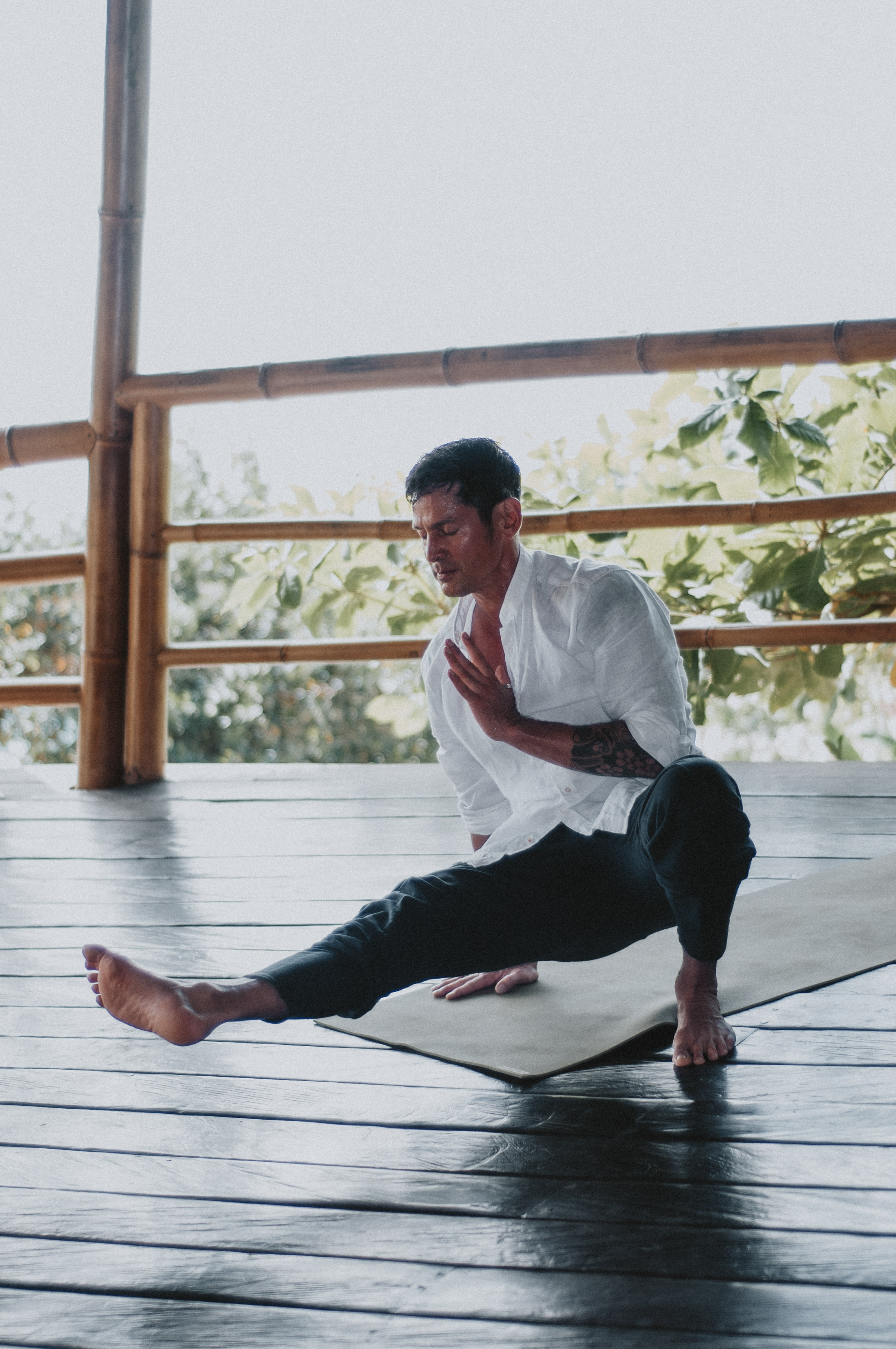 A person practices yoga on a mat in a tranquil, open-air setting surrounded by lush greenery, wearing a white shirt and dark pants, focusing intently on their pose.