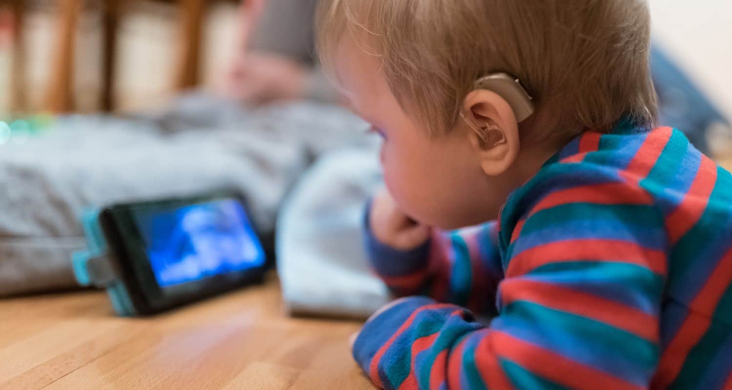 A woman fitting a young girl's hearing aid while the girl smiles, both seated in a cozy setting with plants in the background.