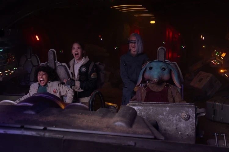 Four young individuals, including an alien with large ears, are joyfully piloting a spaceship. The cockpit is dimly lit, conveying excitement and adventure.