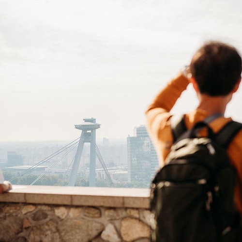 Osoba s batohem zírající na futuristickou věž mostu a panoráma města z kamenného balkonu.
