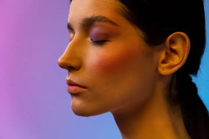 A woman in profile with eyes closed, warm blush across her cheekbone and violet shadow on her lid, against a pink and blue gradient