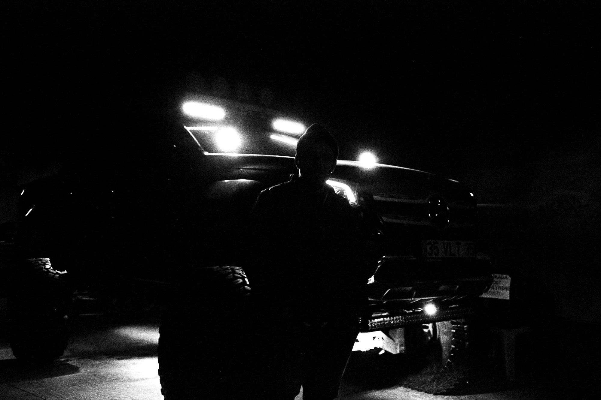 In a dimly lit setting, a person stands in front of a rugged off-road vehicle illuminated by bright LED lights on the roof and front bumper, highlighting its large tires and robust build.