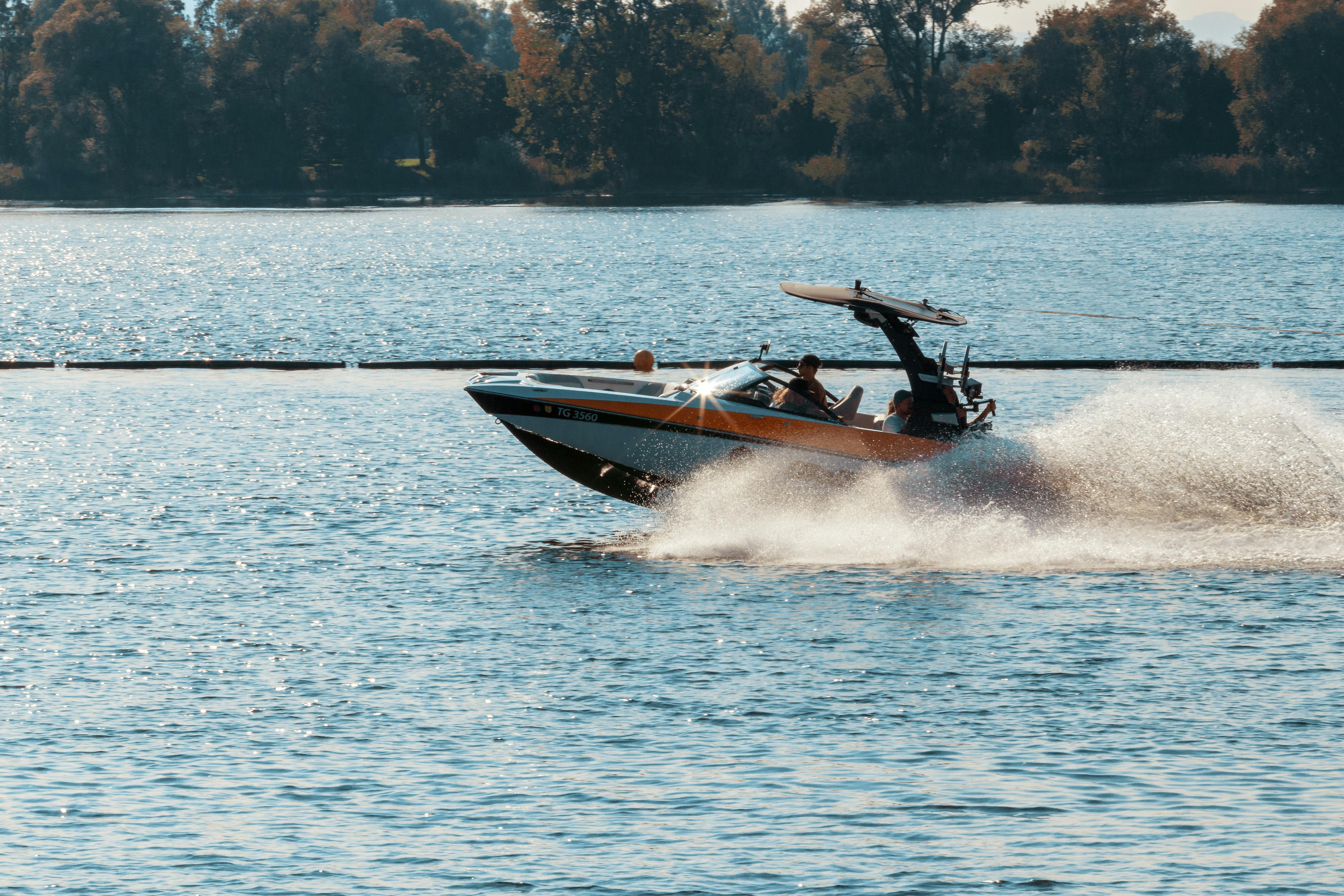 A sleek speedboat skims quickly across a serene lake, leaving a trail of white spray behind, with a backdrop of lush, tree-lined shores under a clear blue sky.
