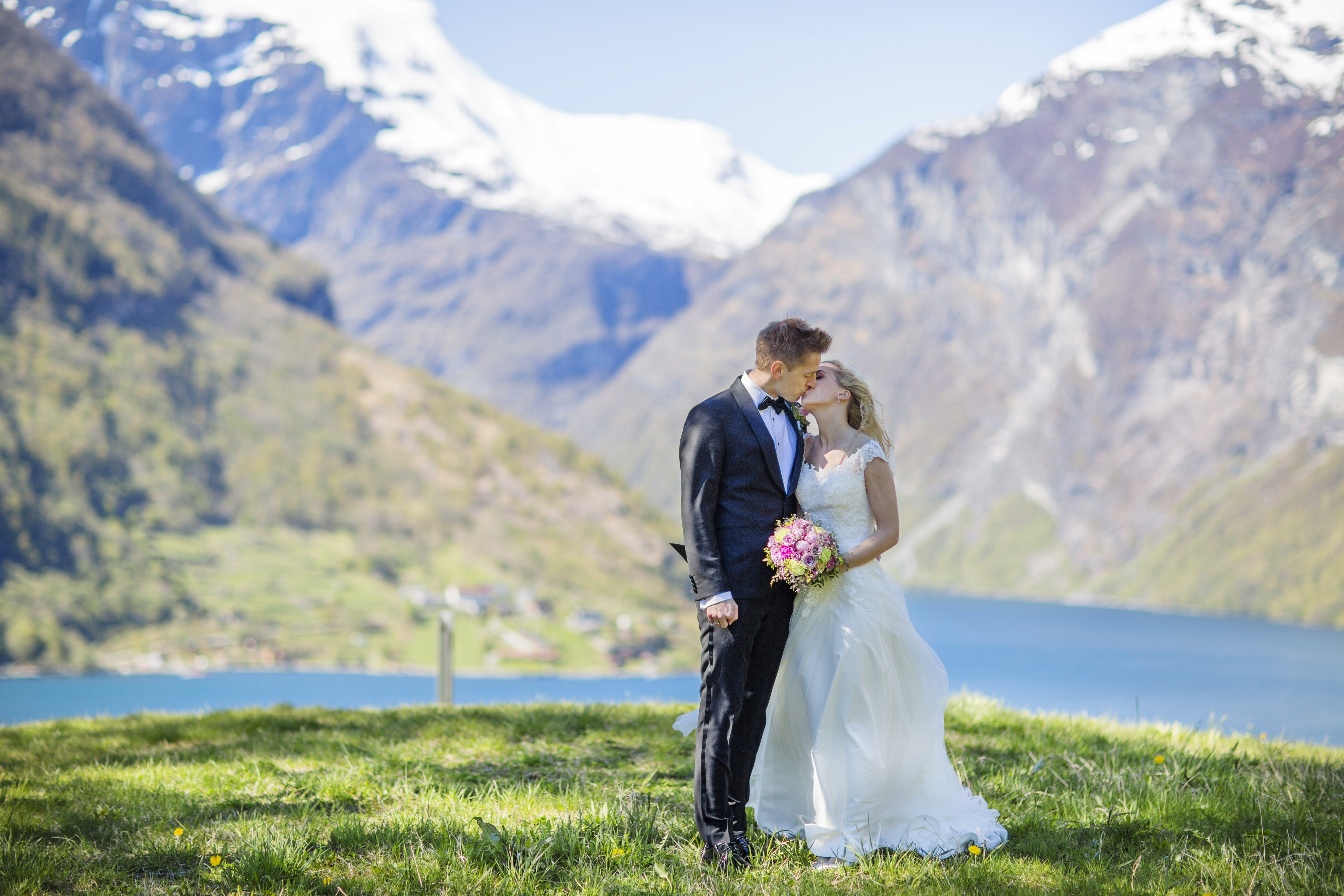 A bride and groom in formal attire stand joyfully under sunlit autumn trees on a cliff edge, overlooking a scenic mountain valley.