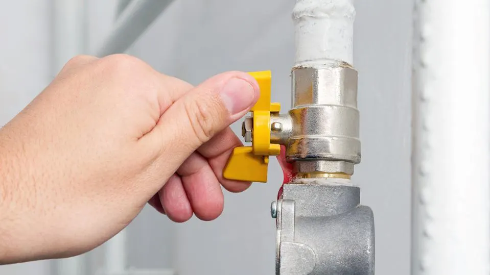 A hand is seen turning a yellow lever on a metal valve connected to a gas piping installation in Asheville. The surroundings are industrial, with parts of the pipe and the valve being visible. The action appears to be regulating the flow of a liquid or gas through the pipe.