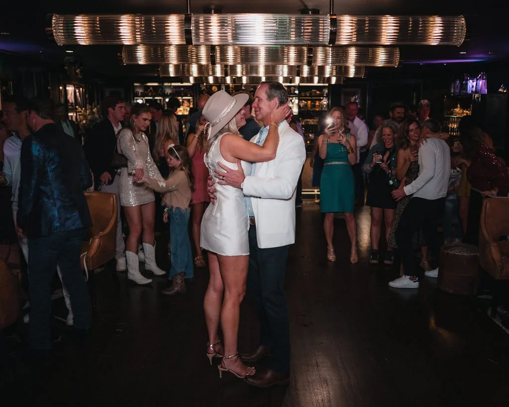 The bride in a short white dress and wide-brimmed hat embraces the groom on the dance floor as guests watch.