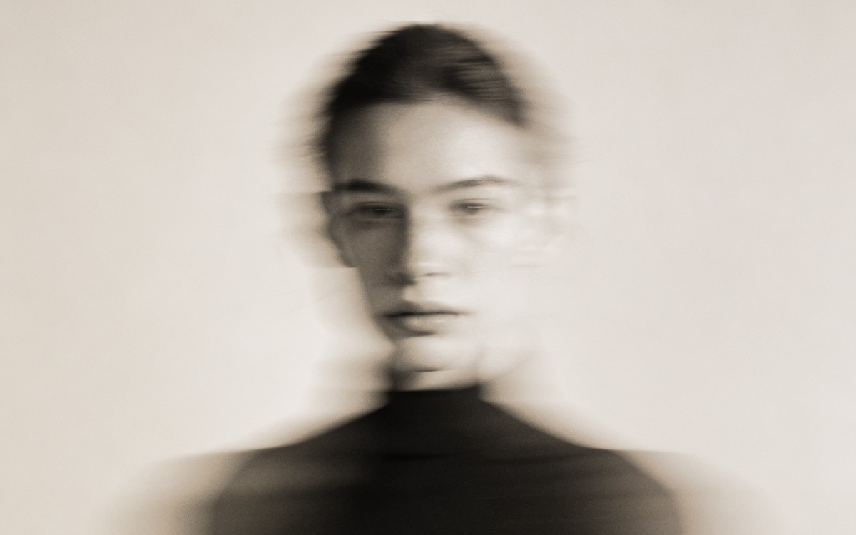 A blurred black-and-white portrait of a person with a neutral expression. The motion effect creates a ghostly, ethereal mood, emphasizing a sense of movement.