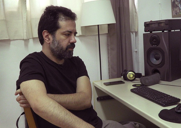 Ο Χάρης Λαμπράκης κάθεται στο στούντιο μουσικής του, πλάι σε ηχεία και μικρόφωνα.