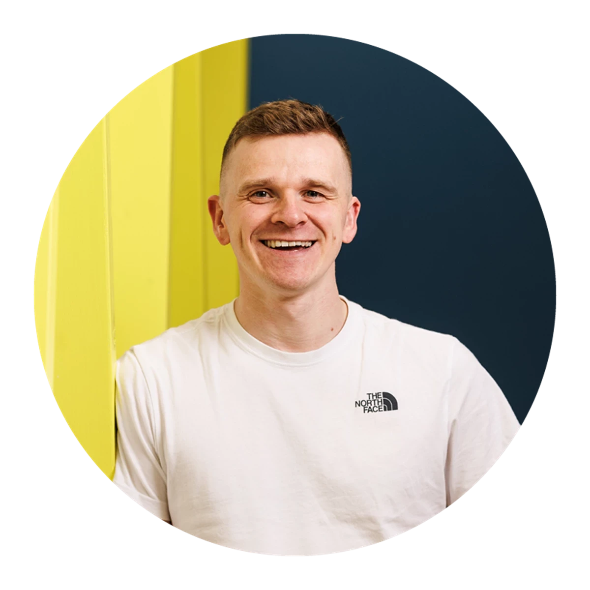 A person wearing a white T‑shirt leans against a bright yellow wall with a dark blue background behind them. The clothing logo is visible on the chest.