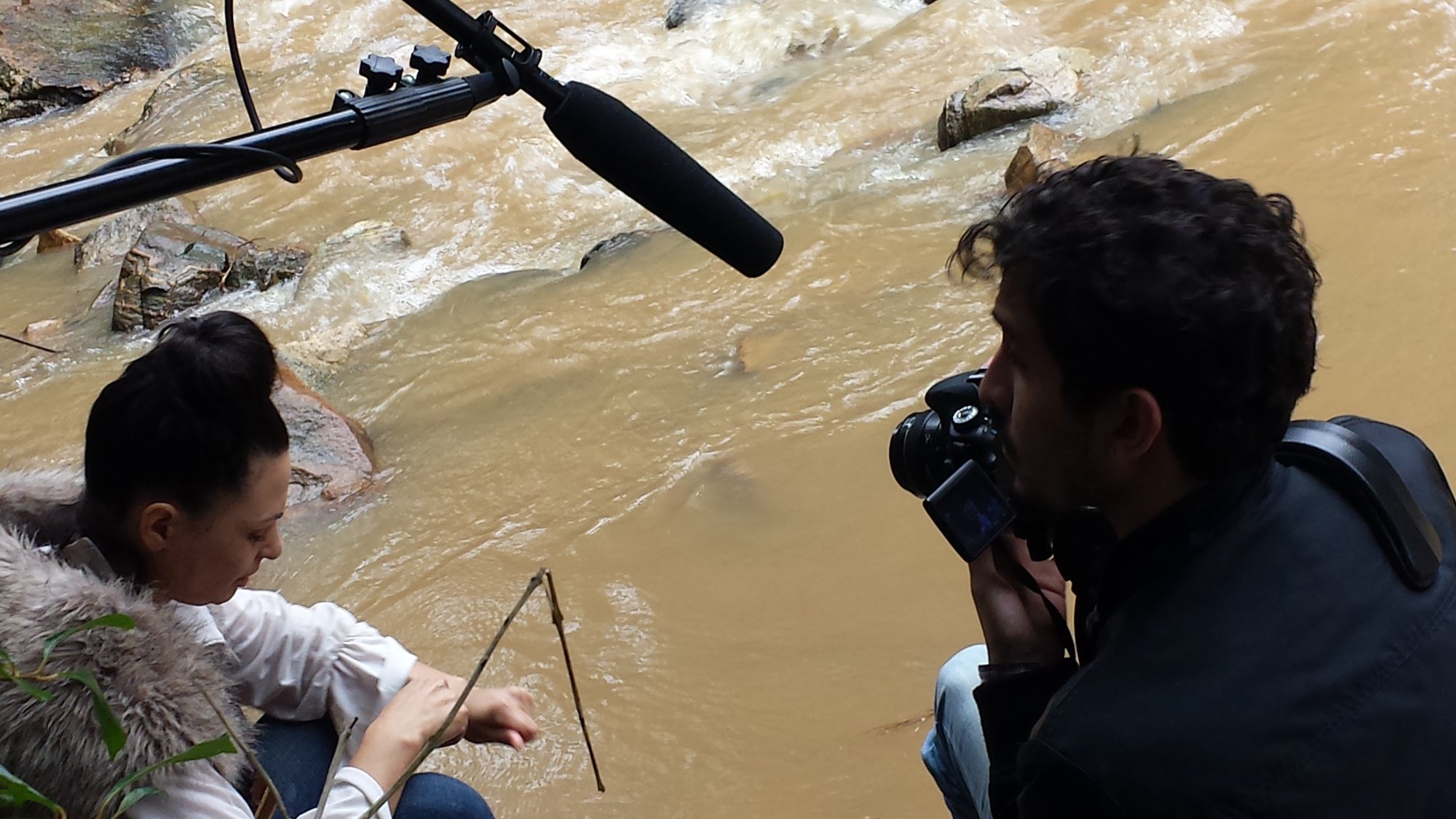 Atriz Vânia Leda sentada à beira do rio barrento, filmada com câmera e microfone de vara