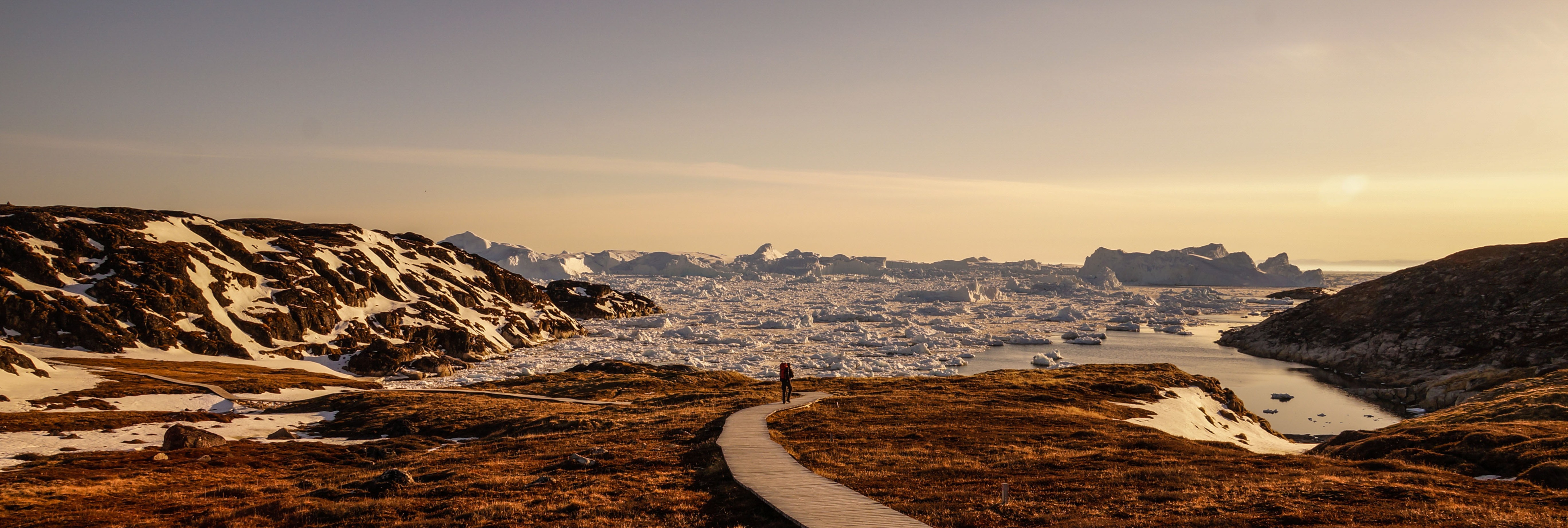 A lone person walking towards an iceberg-filled body of water under the Arctic sun in Greenland
