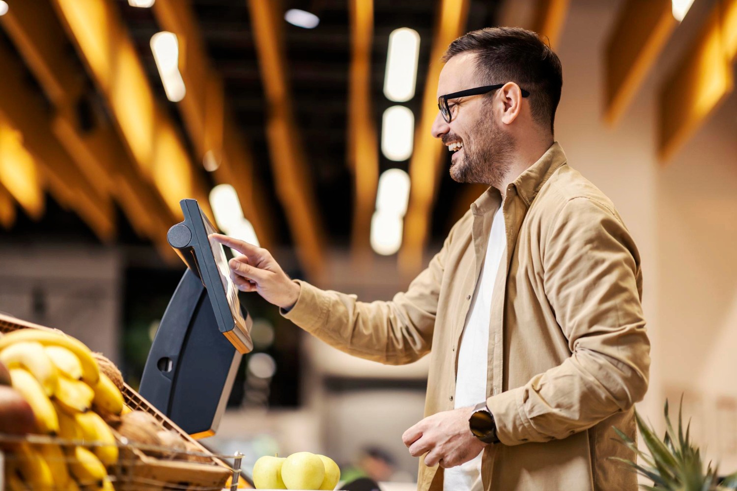 Mann in beiger Jacke bedient Kassenterminal in einem Geschäft mit Obst im Vordergrund.
