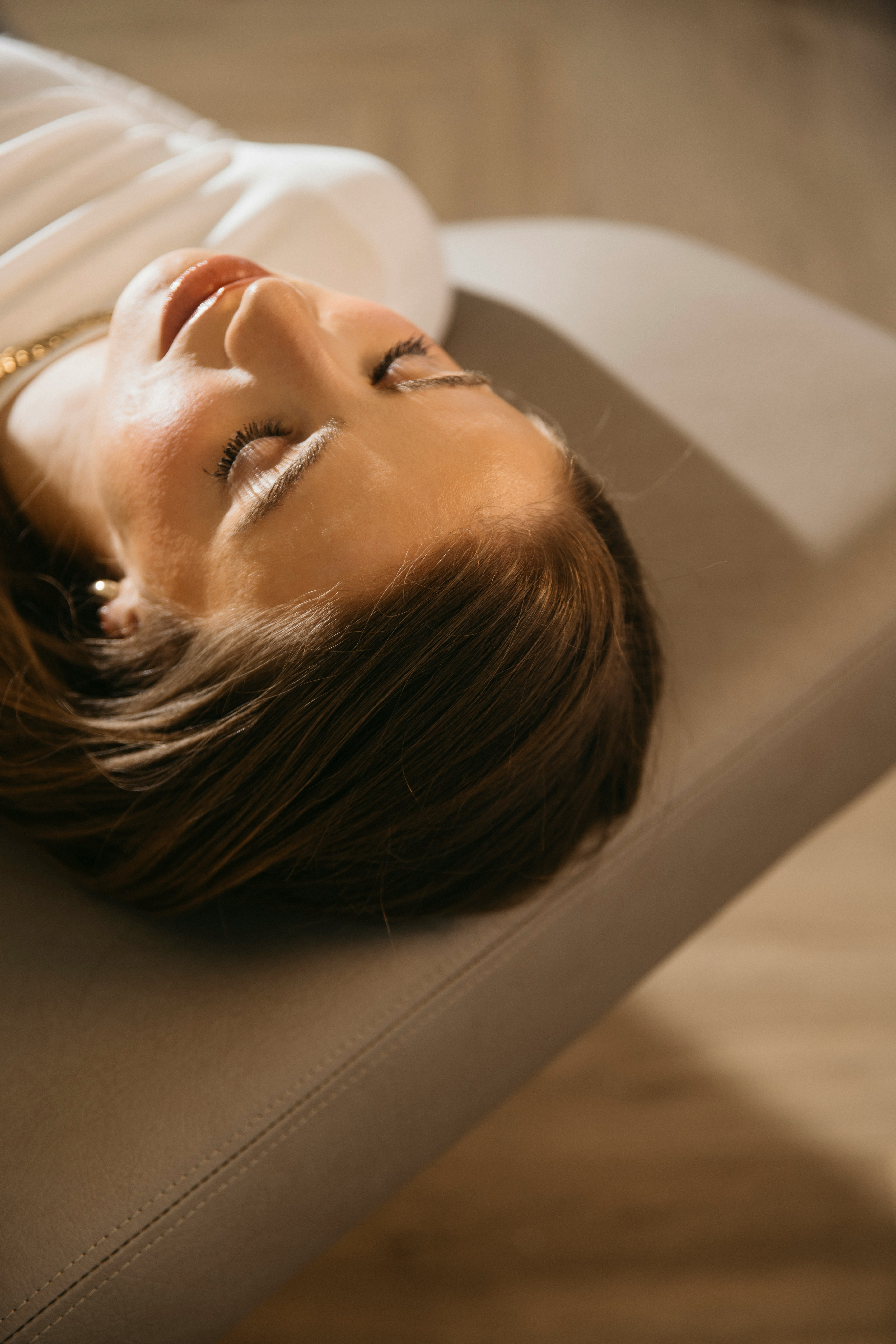 A woman lying on a massage bed, illuminated by soft sunlight streaming through a nearby window. A lush green plant in the background enhances the peaceful and rejuvenating ambiance.