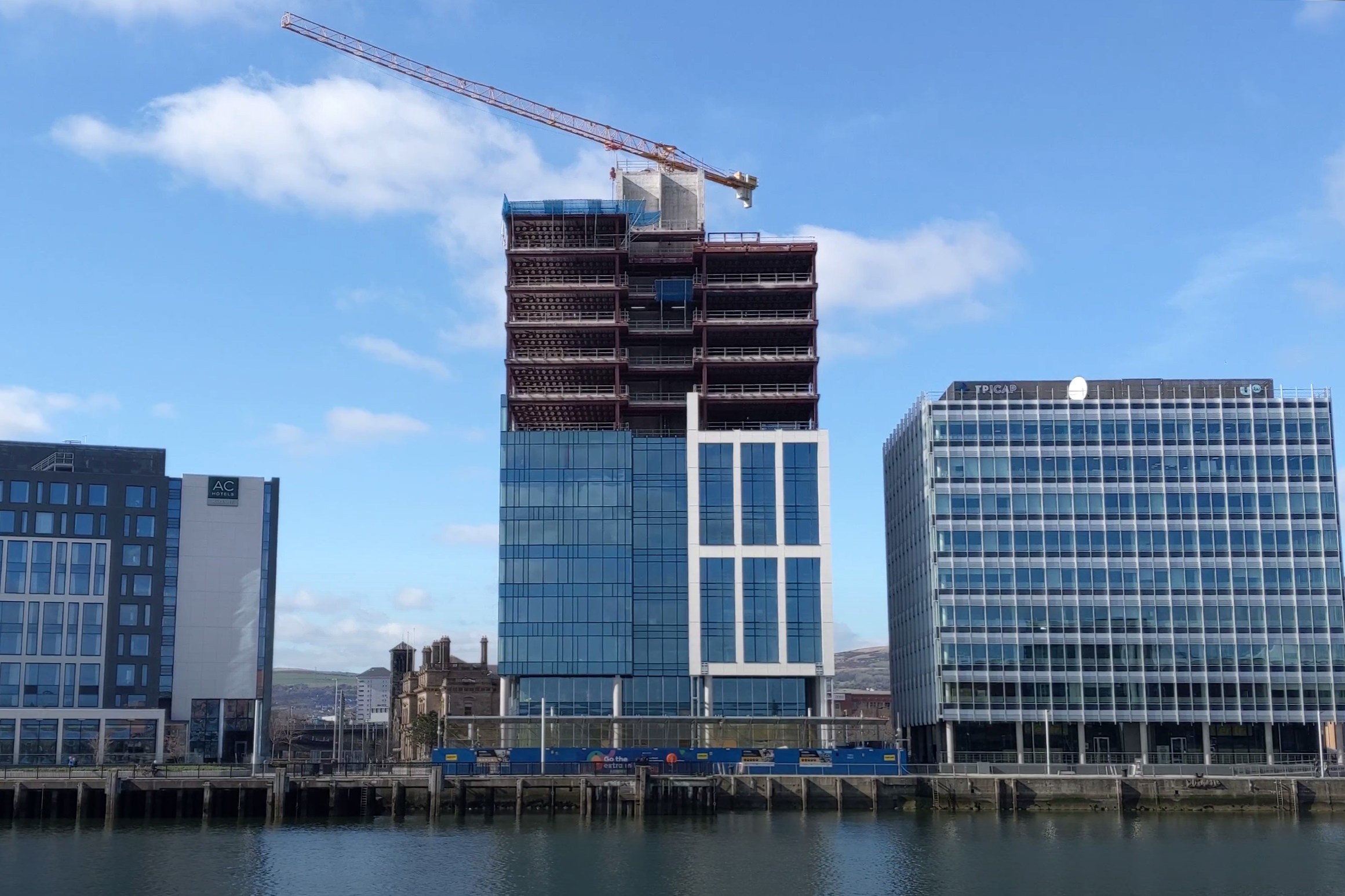 Modern office buildings under construction beside a waterfront with a tower crane overhead.