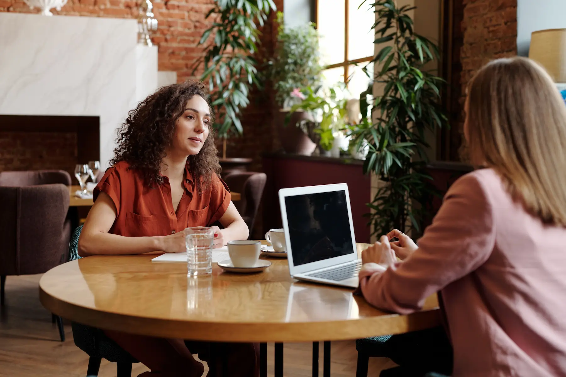 Two people sit at a wooden table in a cozy, brick-walled coffee shop, engaged in conversation, with a laptop and coffee cups in front of them.