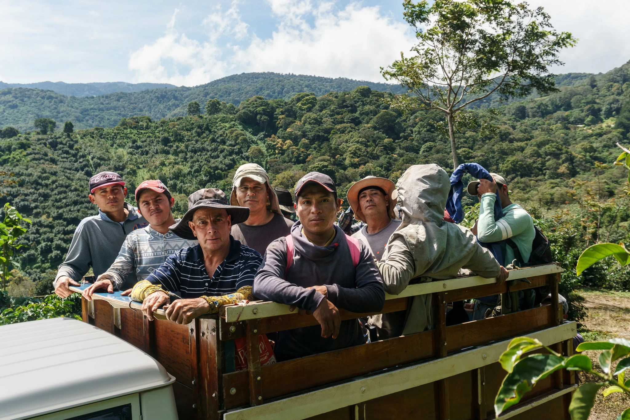 A group of people standing on the back of a truck in a lush, mountainous landscape. They are dressed casually, and some are wearing hats. The surrounding area is green with trees and hills in the background under a partially cloudy sky.