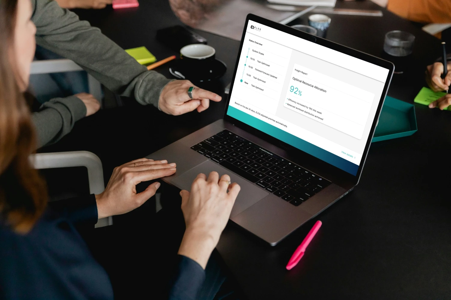 Mehrere Personen sitzen an einem Tisch und betrachten das ELOQ Desktop Interface auf einem Laptop. Das Dashboard zeigt einen Status-Überblick und eine Analyse zur optimalen Ressourcenzuweisung mit einem Effizienzwert von 92 Prozent.