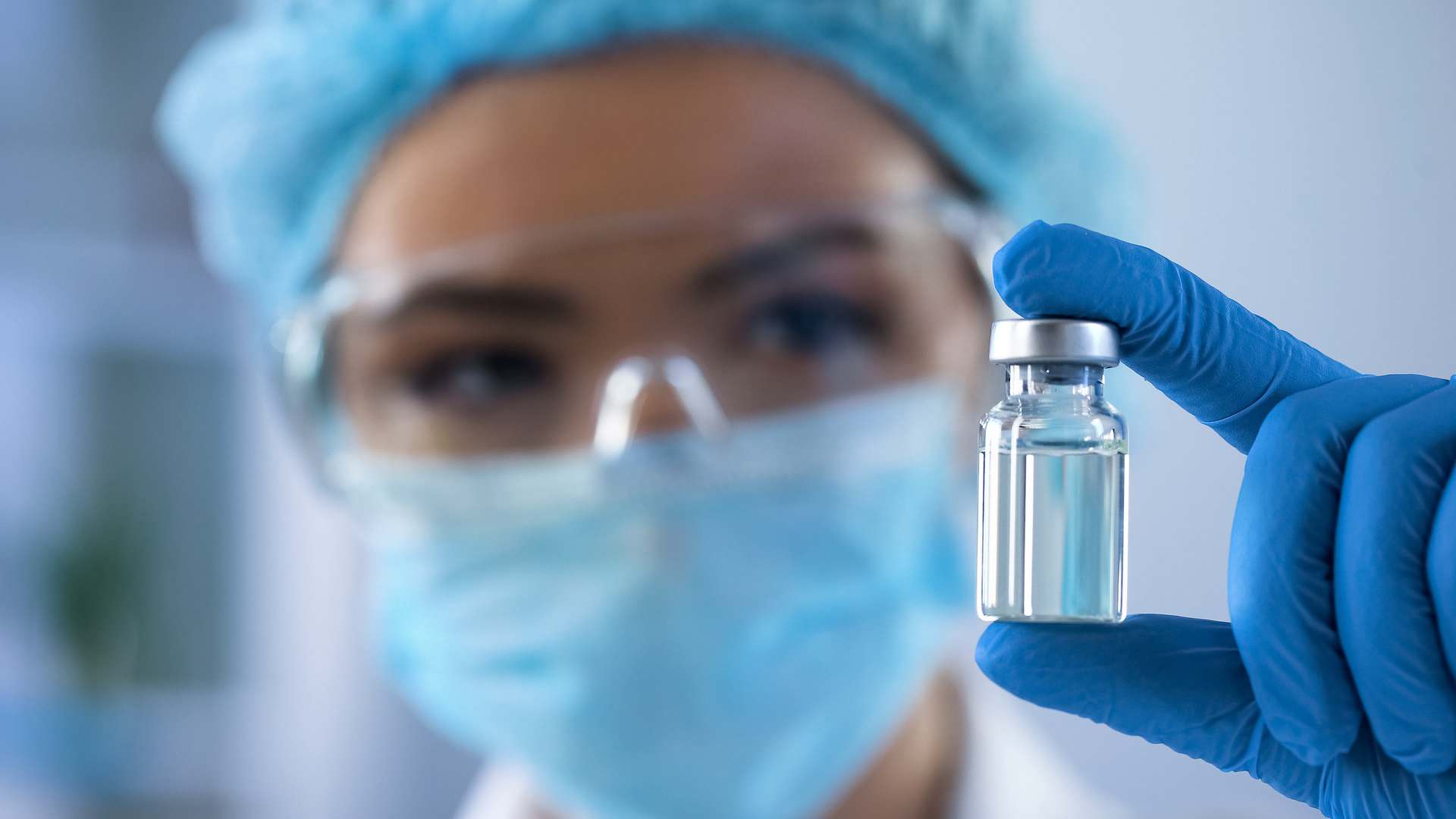 Scientist in a blue mask, hairnet, safety glasses and gloves holding up a small clear vial in a laboratory