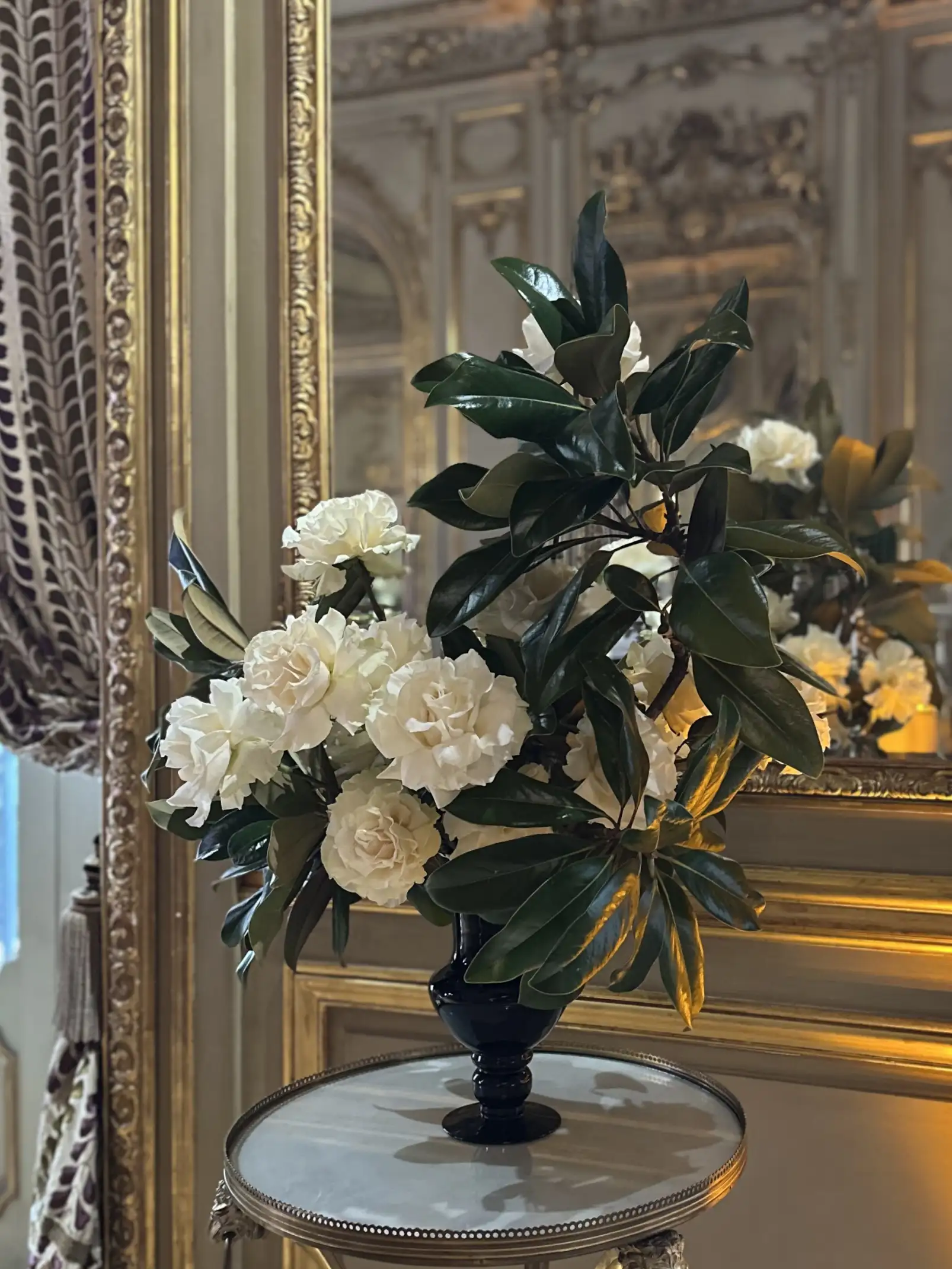 Arreglo floral elegante con flores blancas y hojas verdes brillantes en un jarrón negro sobre una mesa redonda, frente a un espejo con molduras doradas en un entorno de estilo clásico y refinado.