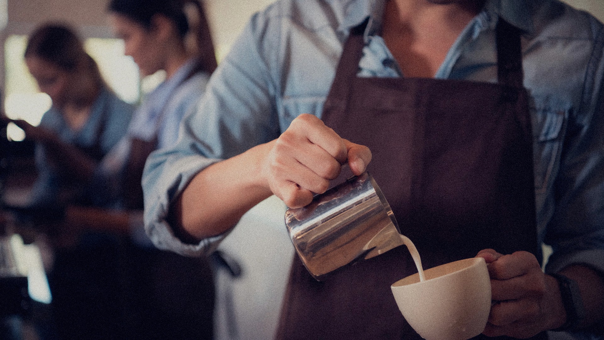 A grainy image of 3 baristas, the closest one to the camera is on the right side pouring frothed milk into a cappucino. They are all wearing chambray shirts with a full body brown canvas apron.